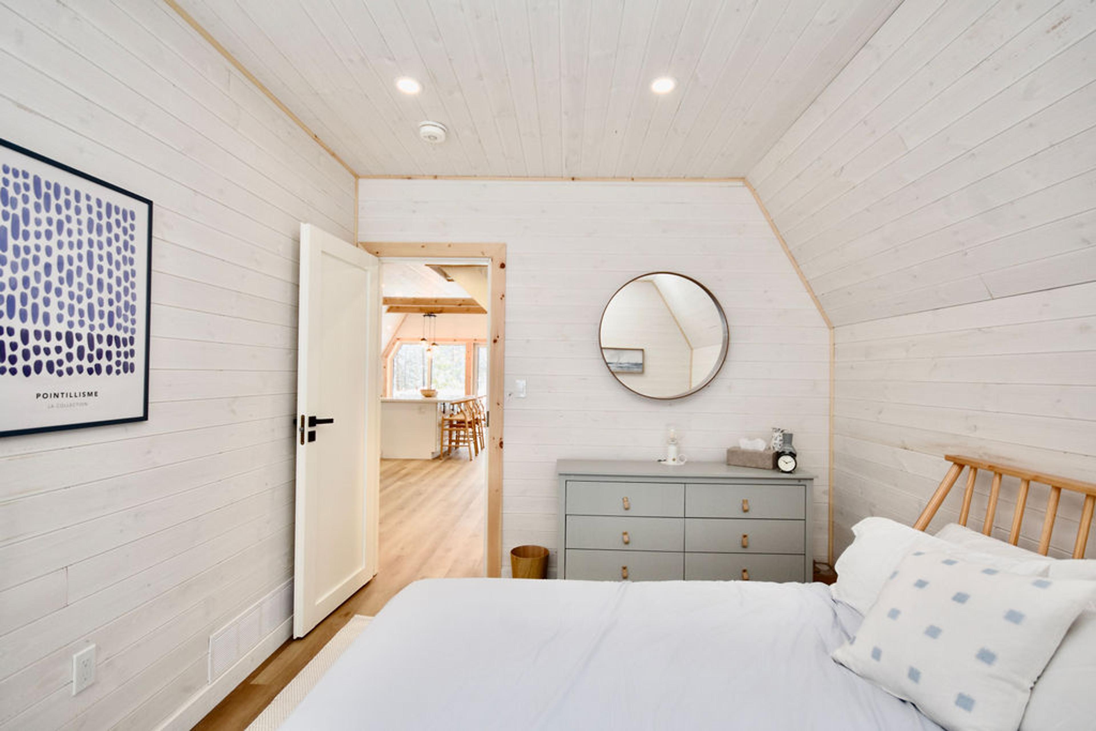 A bright interior view of a modern bedroom inside an A-frame house, featuring whitewashed wood-plank walls and ceiling. A bed with light blue bedding and a patterned throw pillow sits in the foreground next to a sloped ceiling wall. In the center, a grey six-drawer dresser sits below a round wall mirror. An open white door on the left leads into a spacious, sunlit living area with visible kitchen counters and barstools.