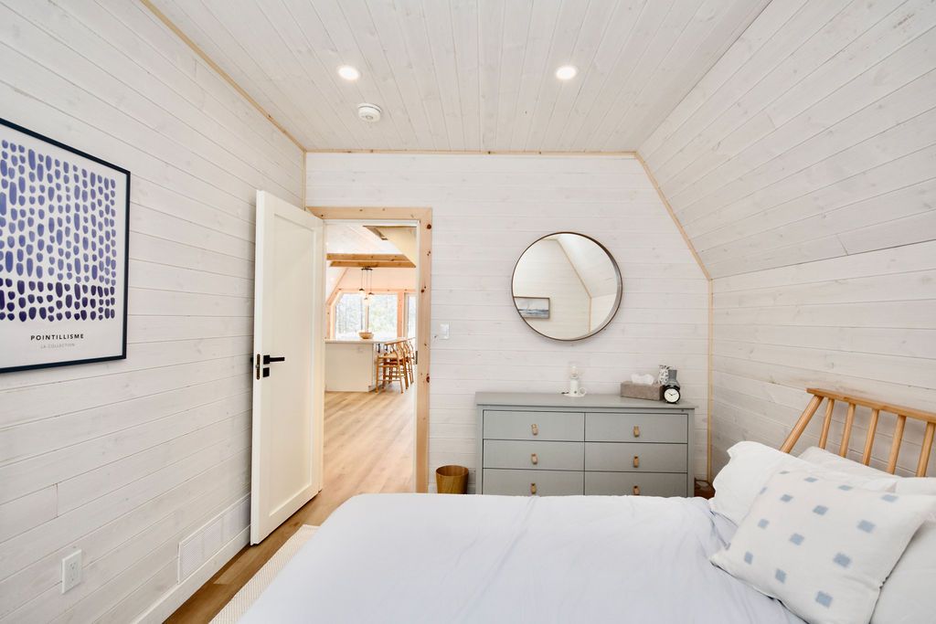 A bright interior view of a modern bedroom inside an A-frame house, featuring whitewashed wood-plank walls and ceiling. A bed with light blue bedding and a patterned throw pillow sits in the foreground next to a sloped ceiling wall. In the center, a grey six-drawer dresser sits below a round wall mirror. An open white door on the left leads into a spacious, sunlit living area with visible kitchen counters and barstools.