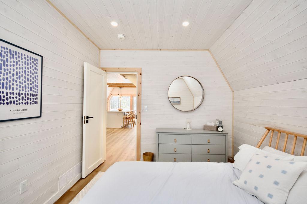 A bright interior view of a modern bedroom inside an A-frame house, featuring whitewashed wood-plank walls and ceiling. A bed with light blue bedding and a patterned throw pillow sits in the foreground next to a sloped ceiling wall. In the center, a grey six-drawer dresser sits below a round wall mirror. An open white door on the left leads into a spacious, sunlit living area with visible kitchen counters and barstools.