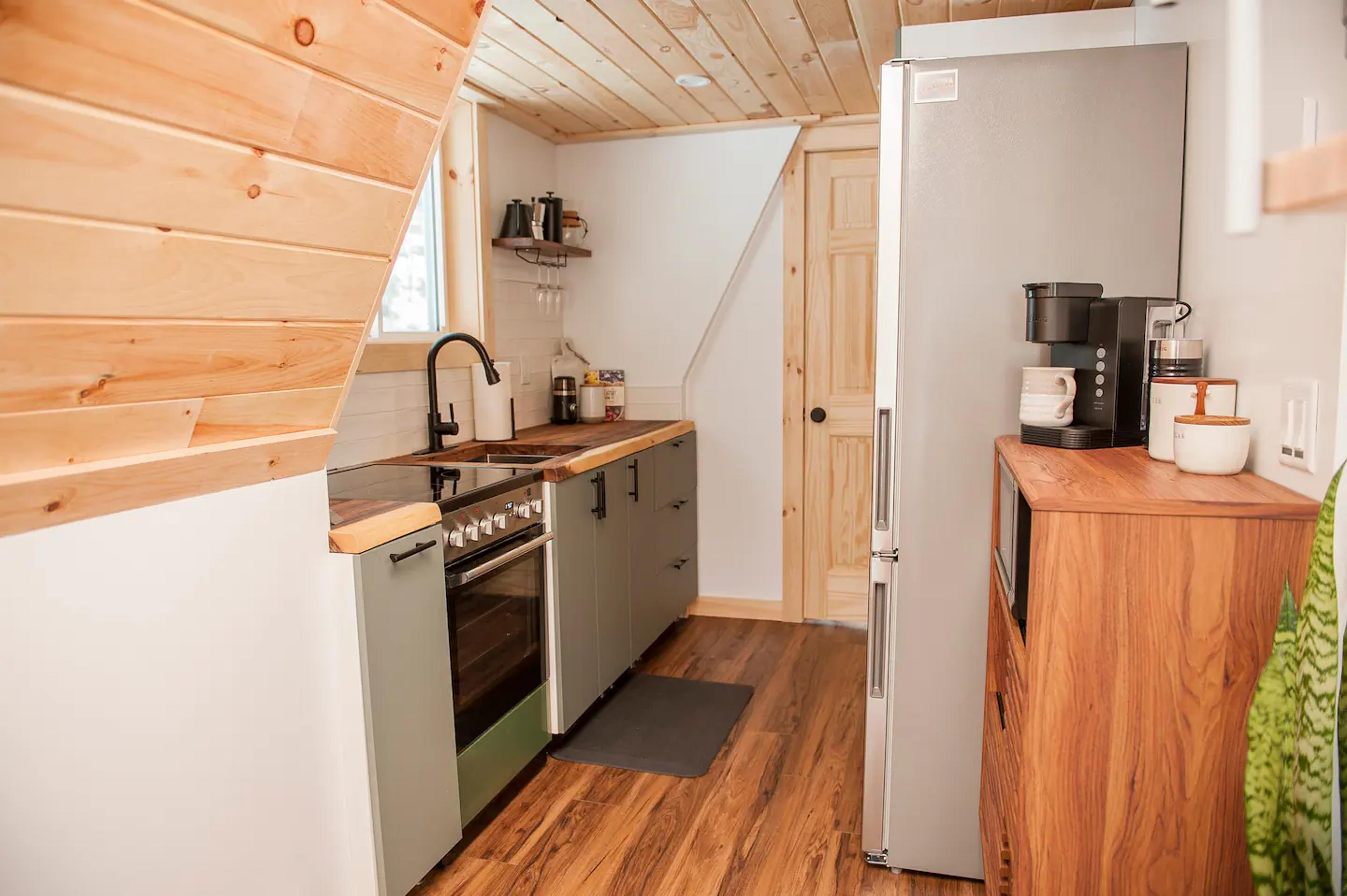 The interior of a compact A-frame kitchen features sage green cabinetry, a live-edge wooden countertop, and a stainless steel refrigerator tucked beneath a light-toned wood-paneled ceiling.