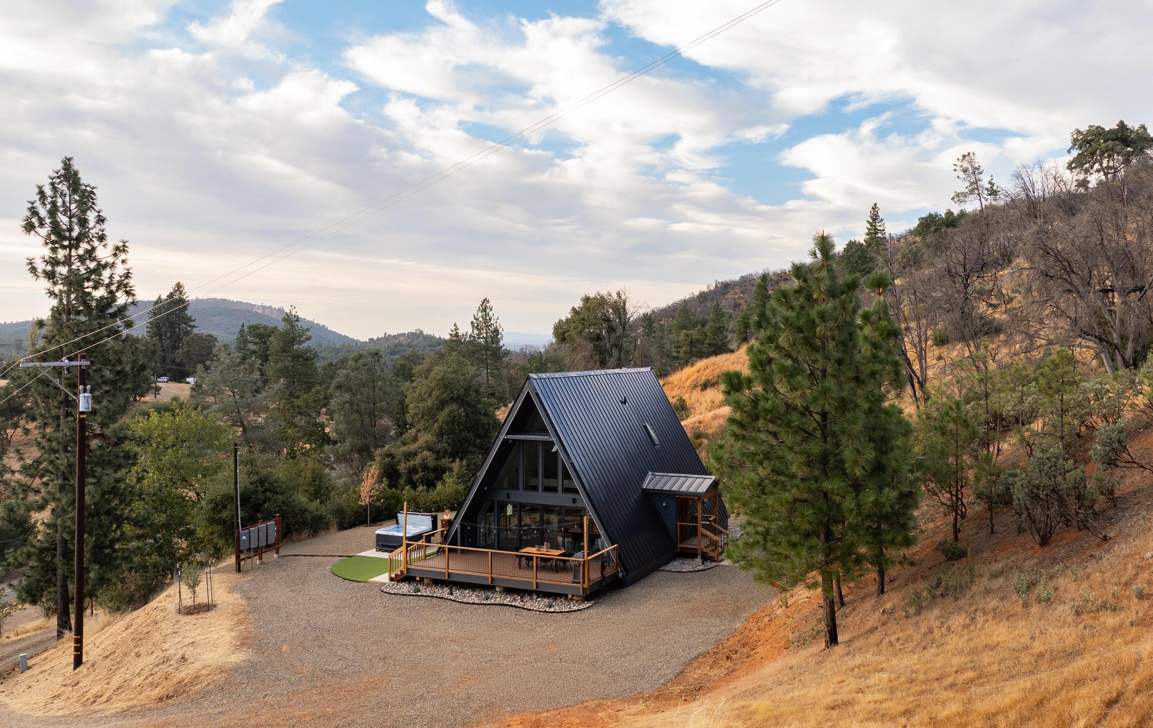 An aerial perspective of a modern A-frame cabin, the "Phoenix Triangle," nestled into a dry, golden-hued hillside. The cabin features a sleek, dark metal roof that extends almost to the ground, large windows on the front facade, and an expansive wooden deck complete with dining furniture and a hot tub. The surrounding landscape consists of scattered pine trees, dry shrubs, and a gravel parking area, all set against a backdrop of rolling hills and a cloudy blue sky.
