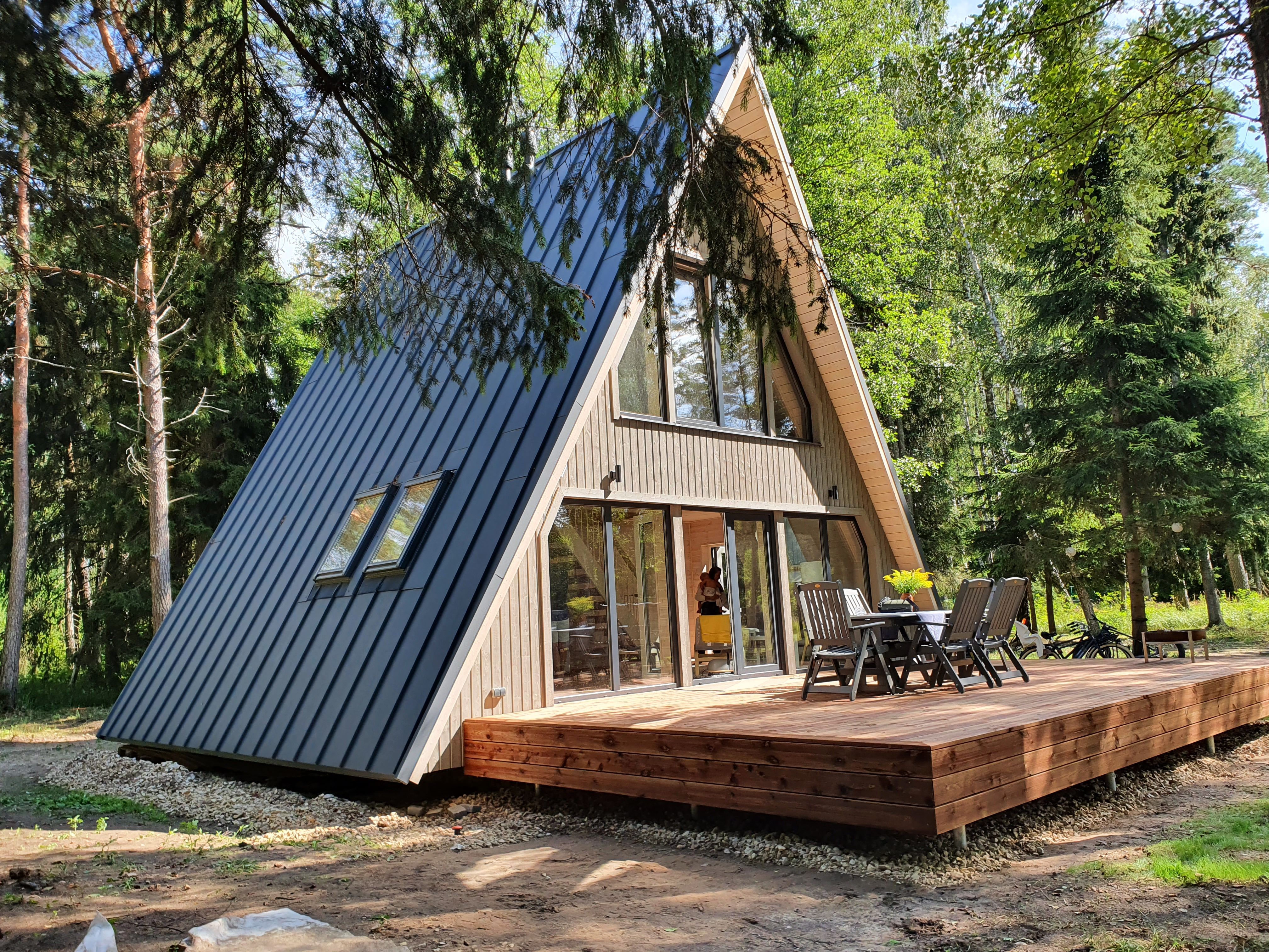 A beautifully finished modern A-frame kit home featuring a sleek dark metal roof and natural wood panels on the facade. The house includes large floor-to-ceiling windows and a spacious wrap-around wooden deck complete with an outdoor dining table and chairs under the shade of mature trees.