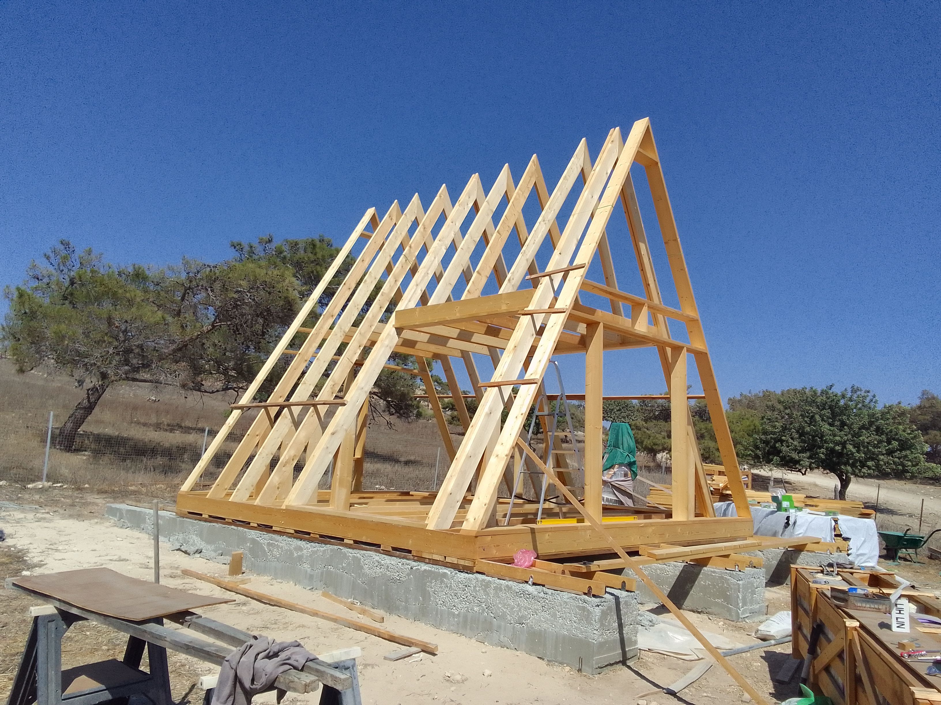 The raw wooden structural framework of an Avrame A-frame kit home raised on a concrete block foundation. The geometric wooden rafters stand sharply against a clear blue sky in an open, arid field, highlighting the precision engineering of the initial assembly phase.