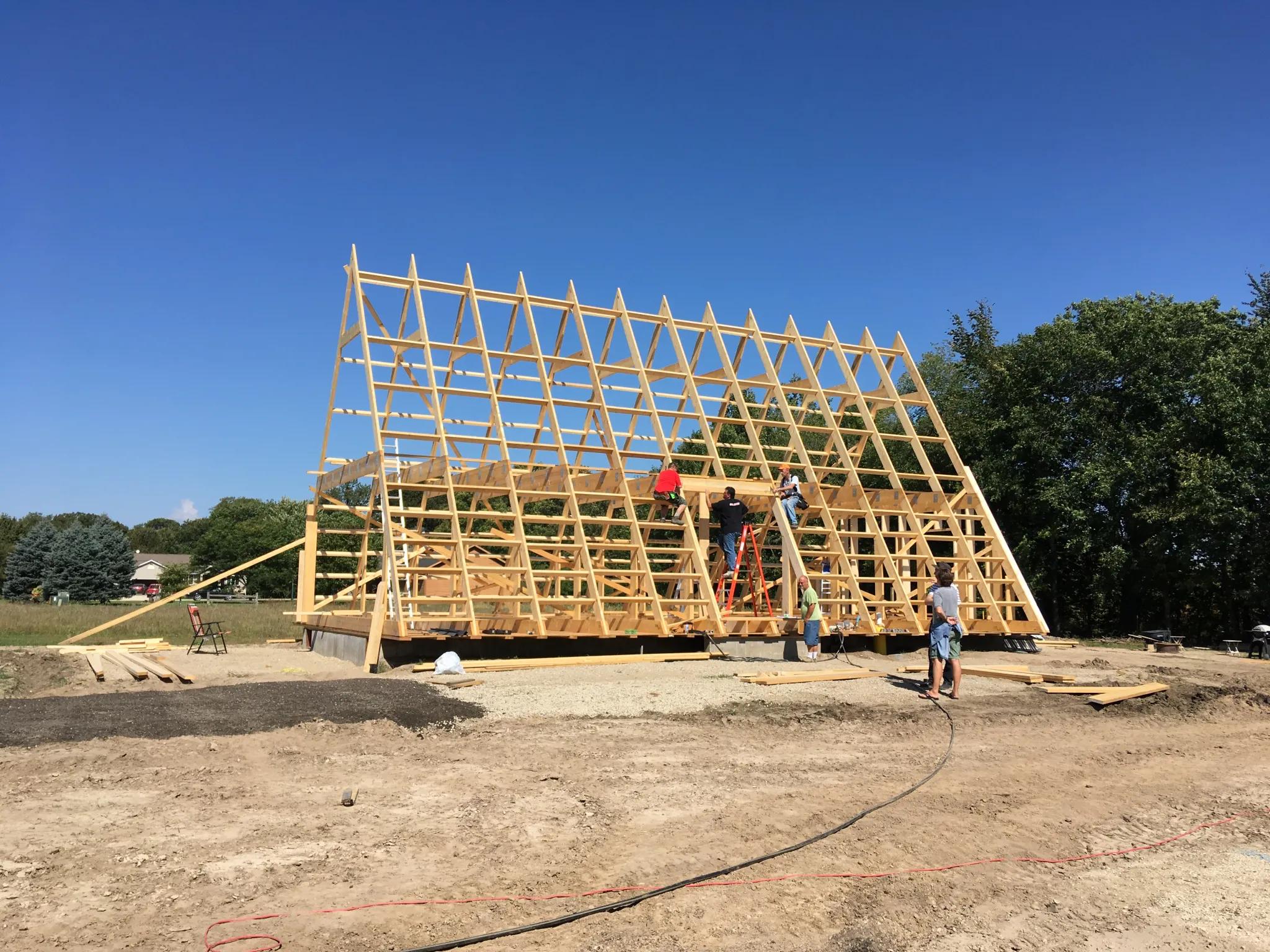 A wide shot shows several construction workers on ladders and the second-story joists of a large, wooden A-frame skeletal structure under a clear blue sky.