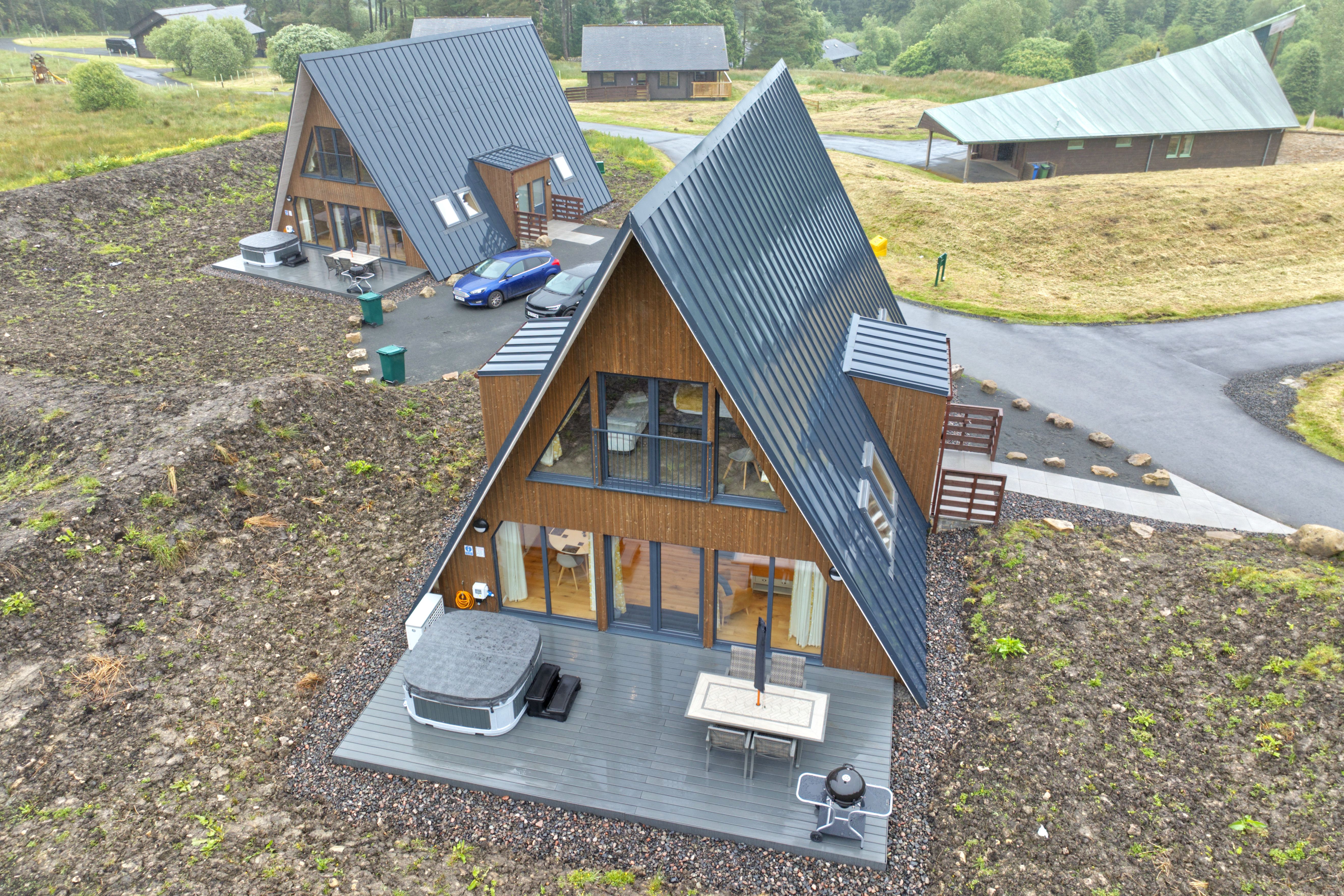 High-angle aerial view of two modern two-story A-frame cabins clad in warm wood with dark metal standing-seam roofs and dormer additions. The cabin in the foreground features a grey patio deck equipped with a hot tub, outdoor dining table, and kettle grill, set against a sloped rural landscape with surrounding paved pathways.