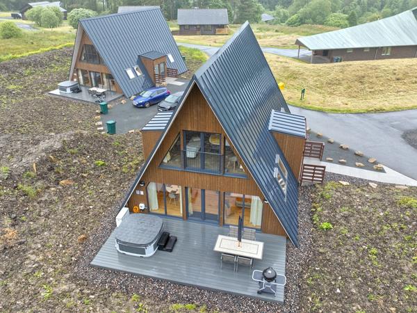 High-angle aerial view of two modern two-story A-frame cabins clad in warm wood with dark metal standing-seam roofs and dormer additions. The cabin in the foreground features a grey patio deck equipped with a hot tub, outdoor dining table, and kettle grill, set against a sloped rural landscape with surrounding paved pathways.