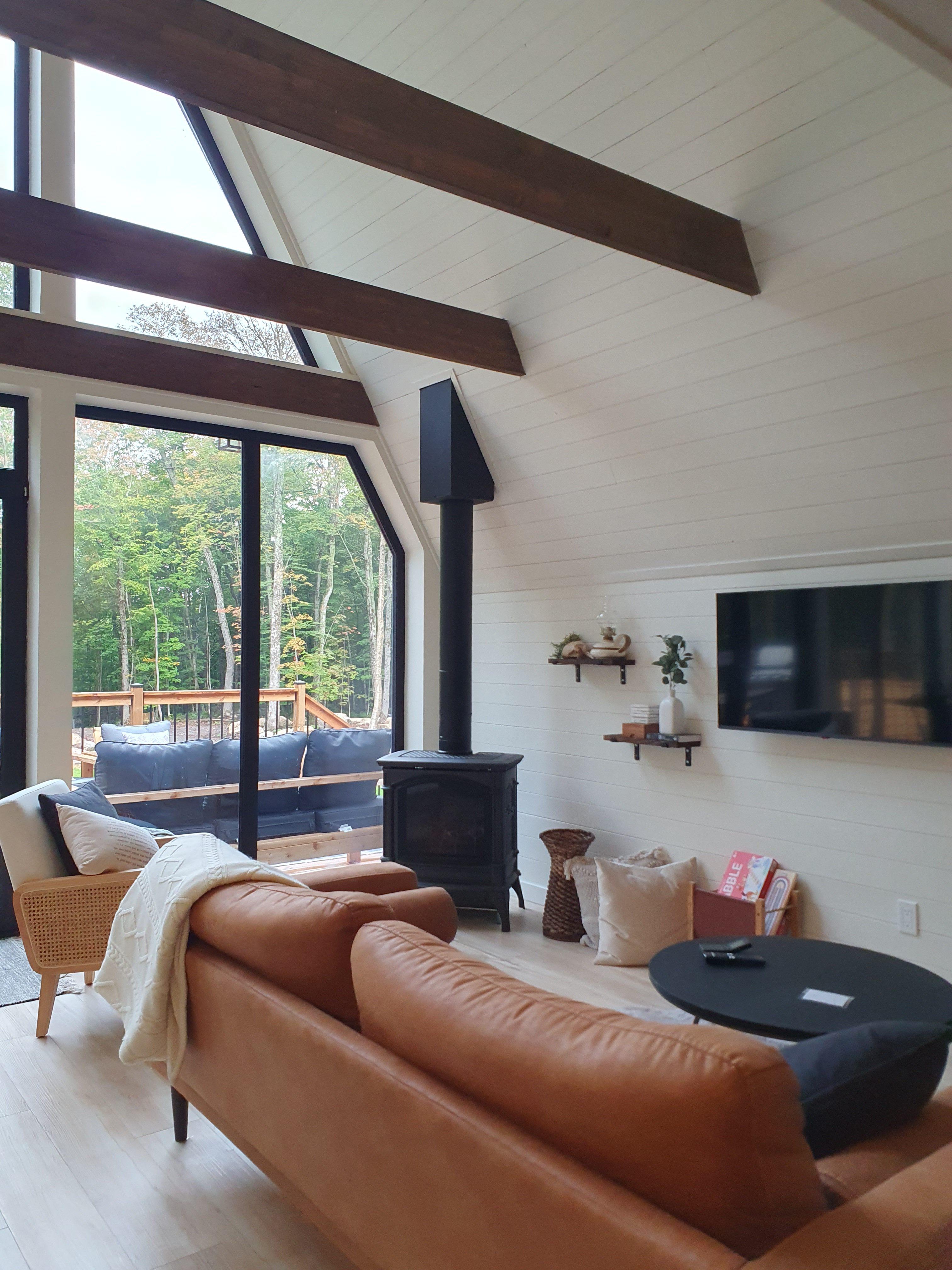 The interior of a modern A-frame living room features a tan leather sofa, a black wood-burning stove, and vaulted ceilings with dark wooden beams that overlook an outdoor deck.