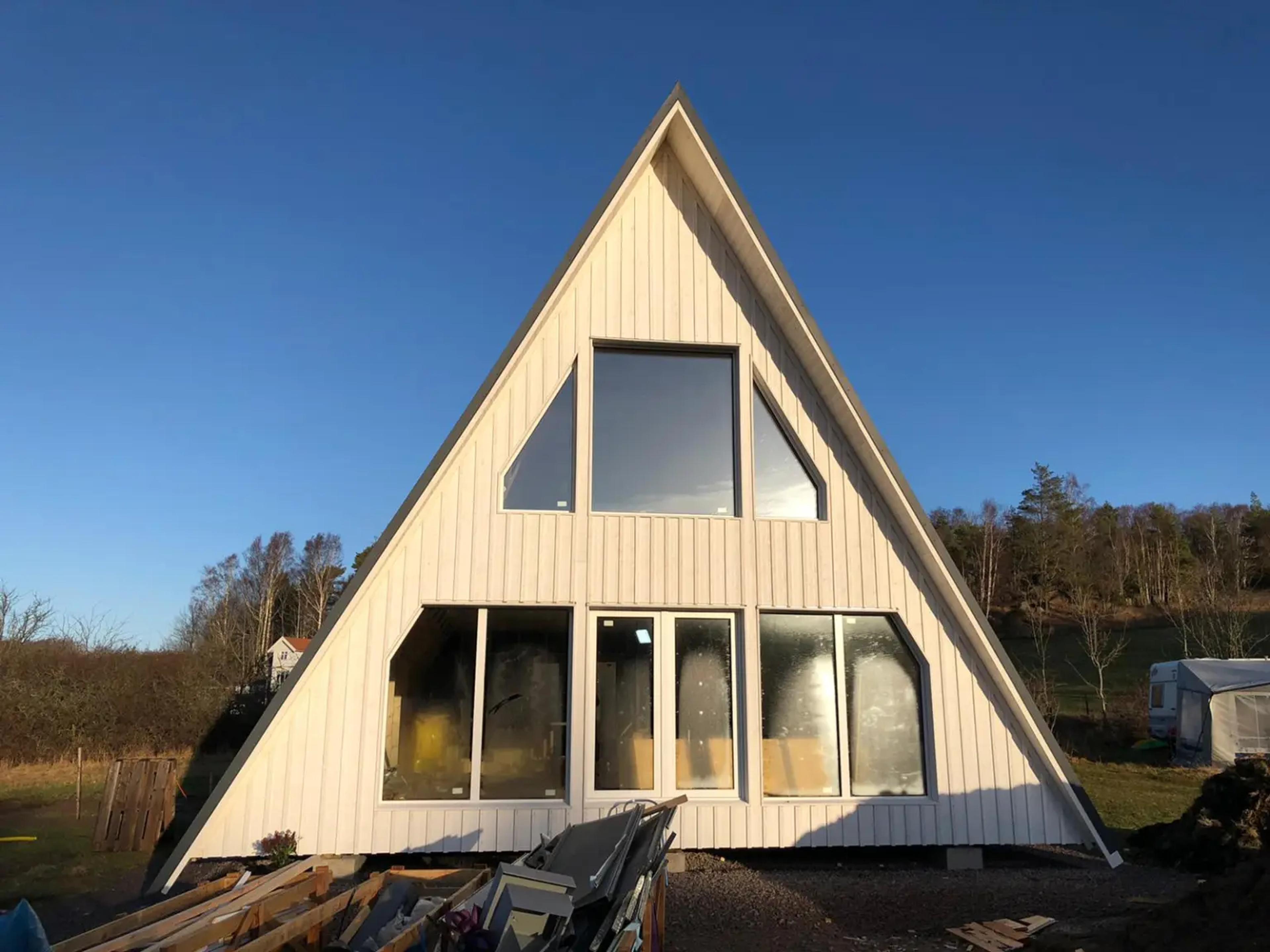 A white A-frame house with vertical siding and large glass windows stands under a clear blue sky on a construction site.