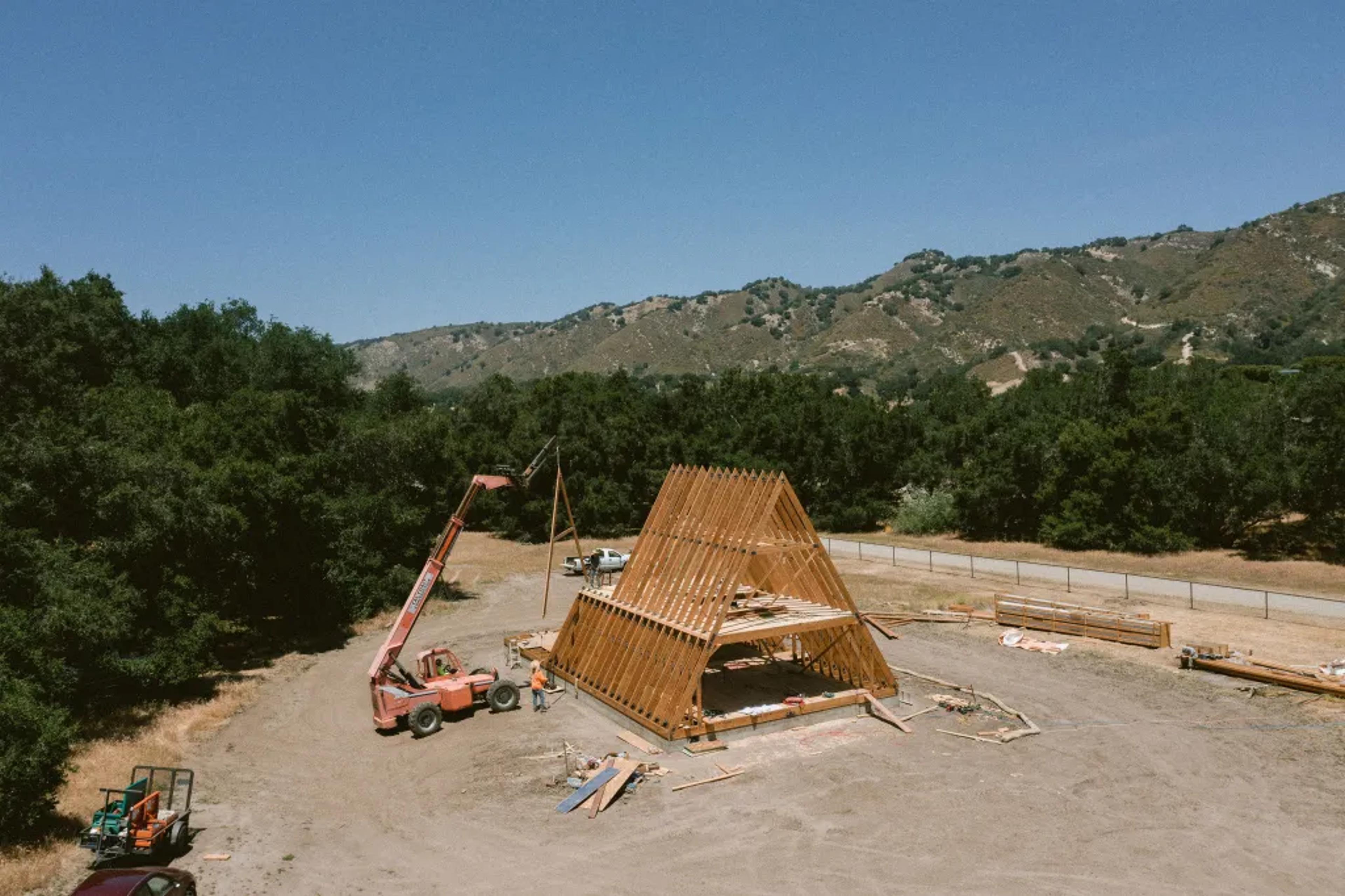 An aerial view shows a telehandler crane assisting workers in the structural assembly of a large timber A-frame house on a dry, open construction site nestled against rolling hills.