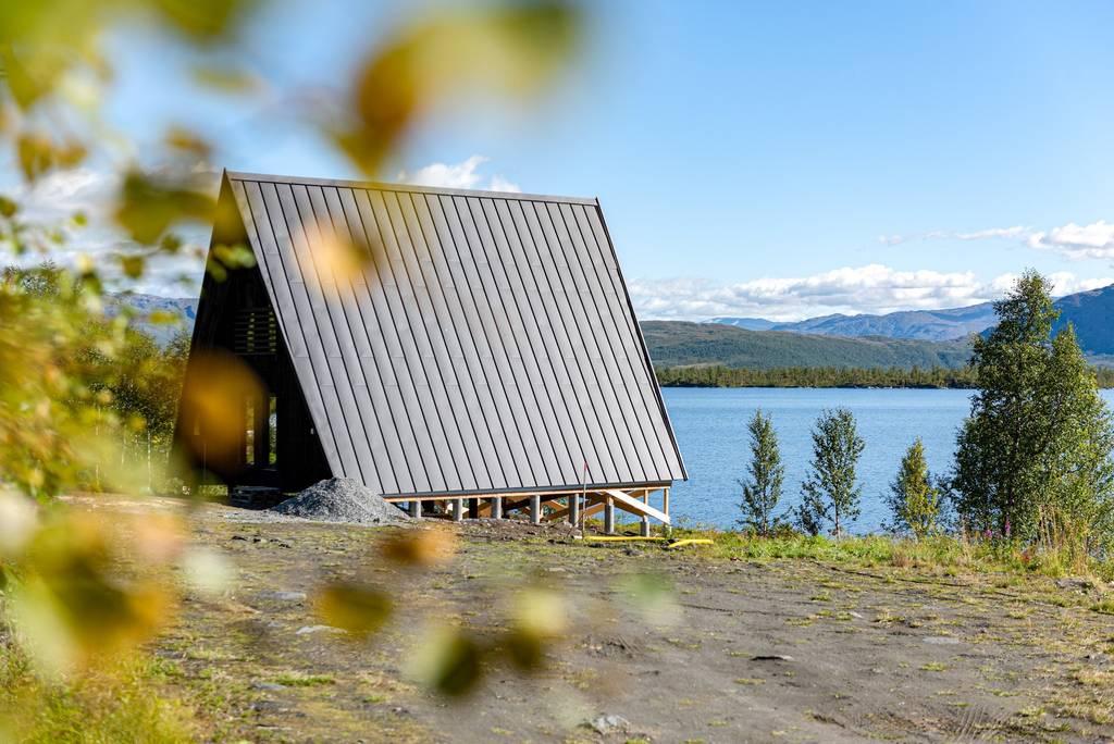 A modern A-frame cabin with dark metal roofing stands on a raised foundation overlooking a serene lake and mountain range.