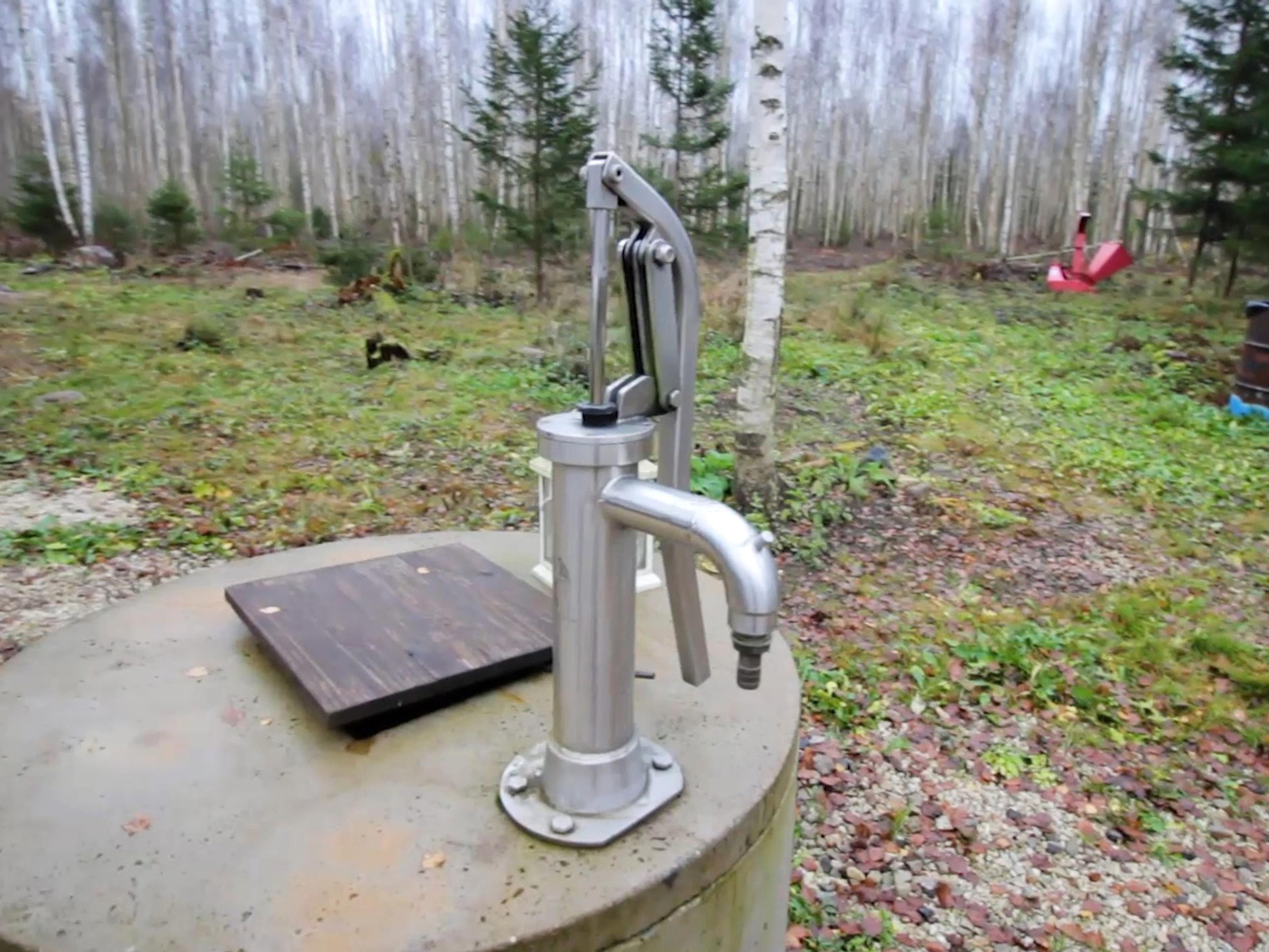A stainless steel manual hand pump is mounted on a concrete well cover in a rural, wooded setting.
