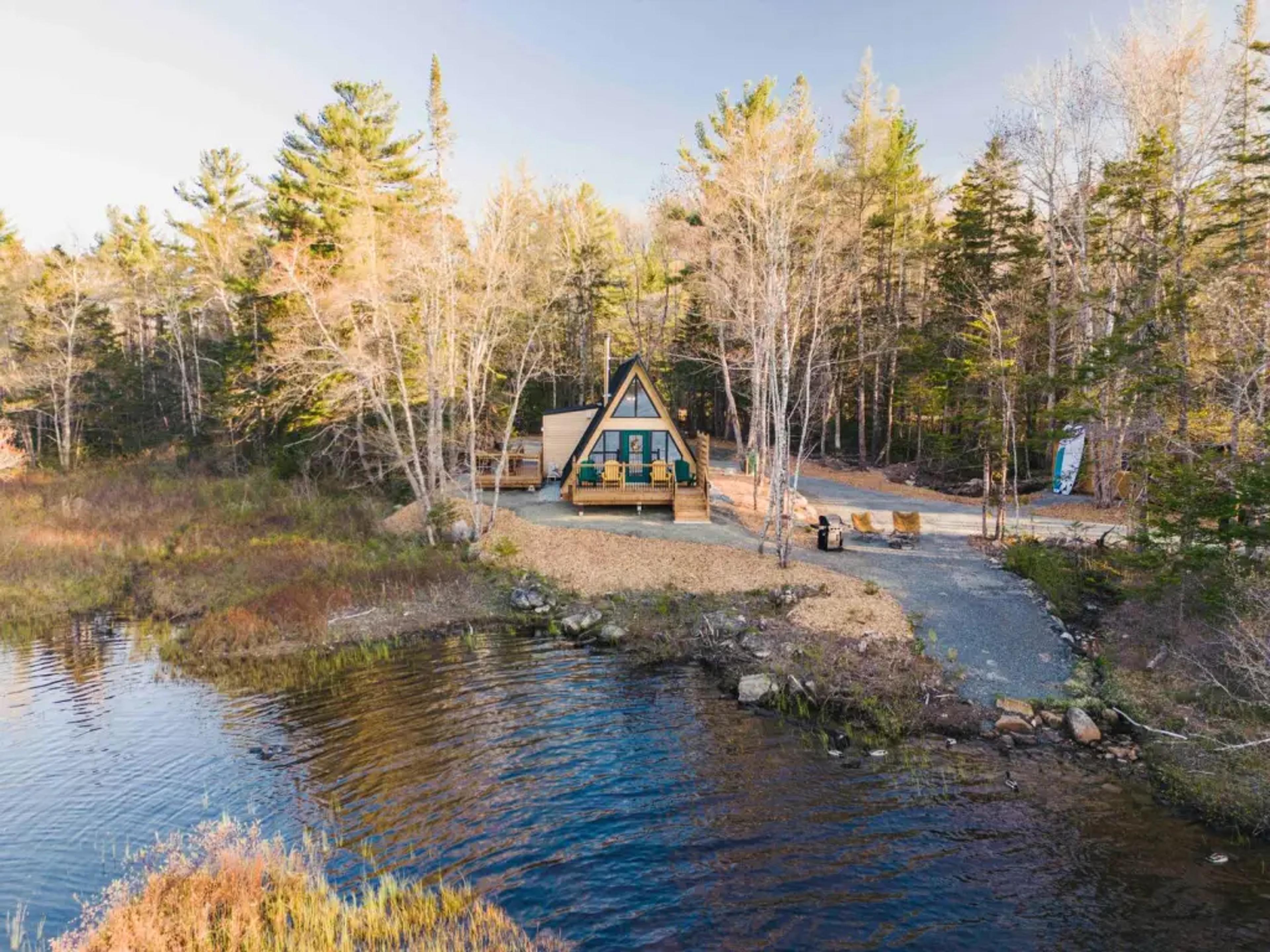 An aerial view shows a cozy A-frame cabin with a large front deck nestled among autumn trees along a winding dirt path near a rippling lake.