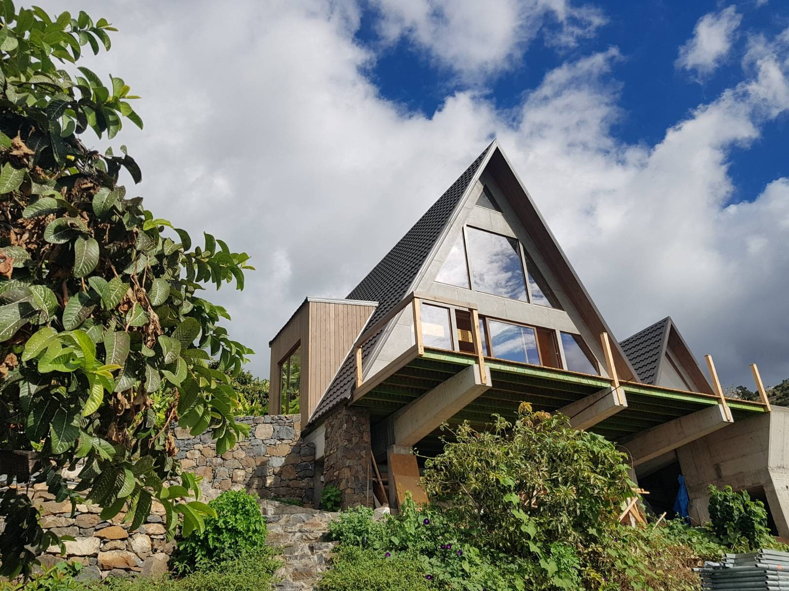 A custom Avrame Trio build on a steep, terraced hillside under a bright, cloudy sky. This image demonstrates how self build kits can adapt to tropical or coastal climates, featuring a large elevated deck and stone-walled landscaping that integrates with the natural terrain.