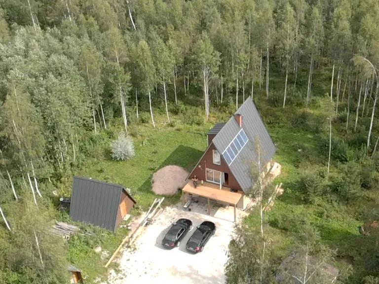 An aerial view shows an off-grid A-frame house equipped with solar panels on its steep metal roof, situated in a forest clearing with a smaller A-frame outbuilding and two cars parked nearby.