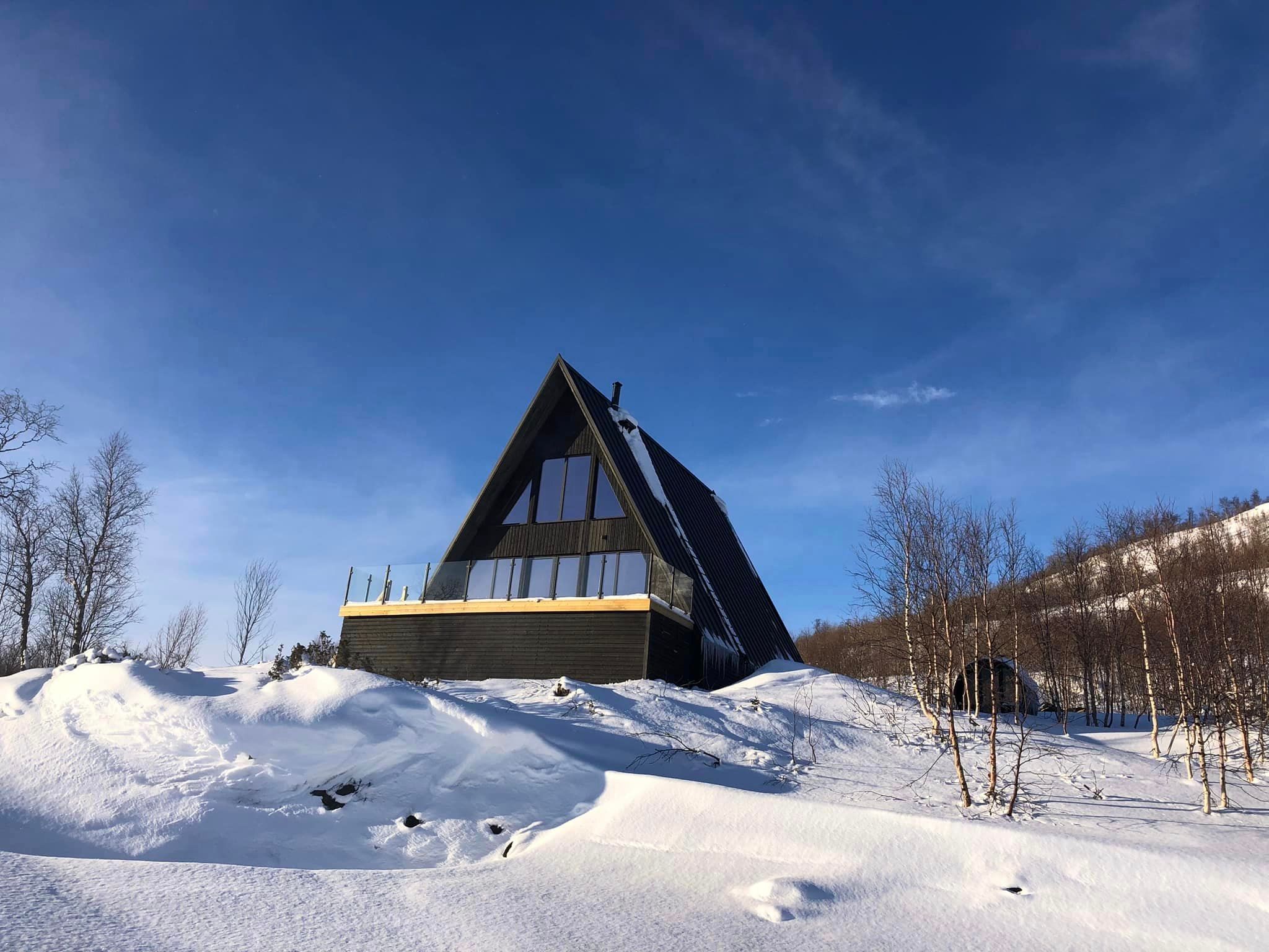 A striking black A-frame cabin perched on a snowy hillside under a clear blue sky. The steep roof design is shown effectively shedding snow, making it a perfect mountain cabin for extreme winter climates where structural strength and heat retention are vital.