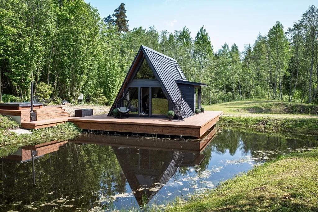 A modern, black A-frame cabin sits on a large wooden cantilevered deck extending over a tranquil pond, perfectly reflecting the structure and the surrounding lush green forest.