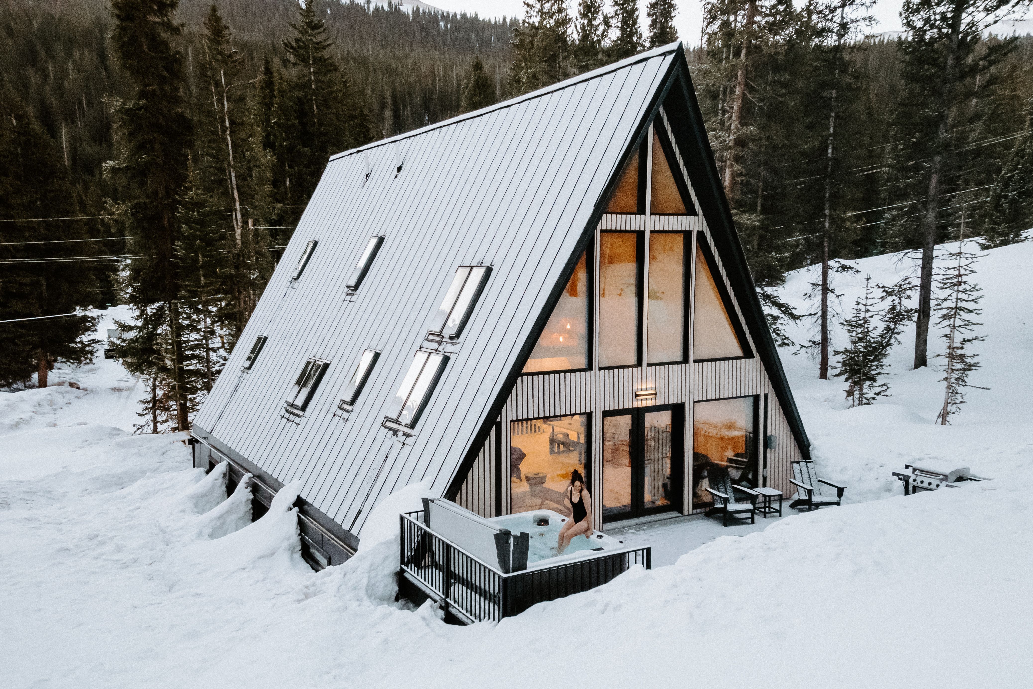 A stunning finished A-frame green home nestled in a snowy mountain landscape. The photo showcases a "tight envelope" design with large energy-efficient windows, a modern metal roof, and a luxury outdoor hot tub, proving that a smart house construction project can result in a high-end, cozy sanctuary.