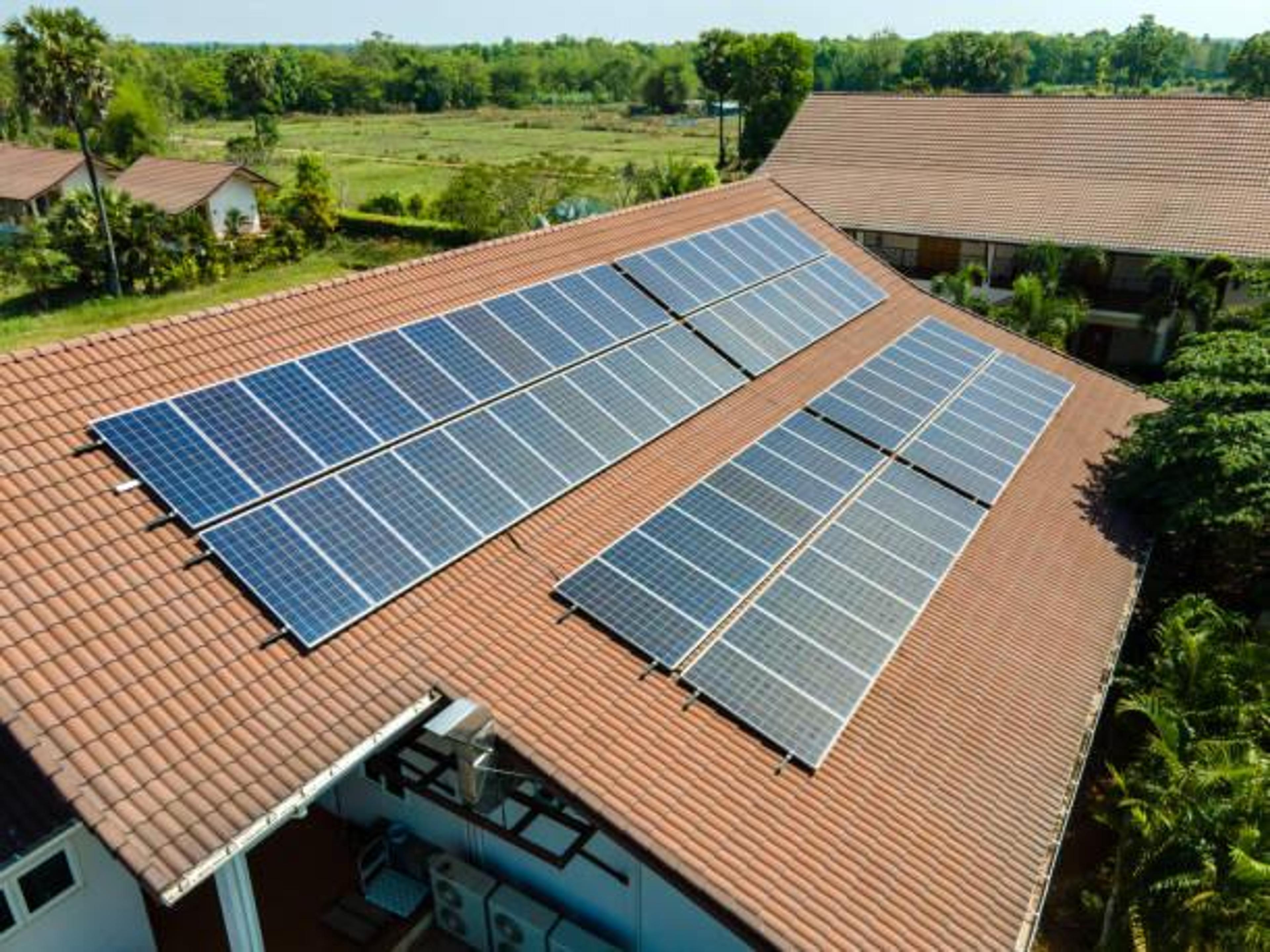 An aerial view shows multiple solar panels installed across the brown tiled roof of a building, overlooking a lush green landscape.