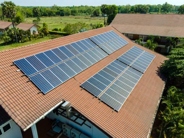 An aerial view shows multiple solar panels installed across the brown tiled roof of a building, overlooking a lush green landscape.