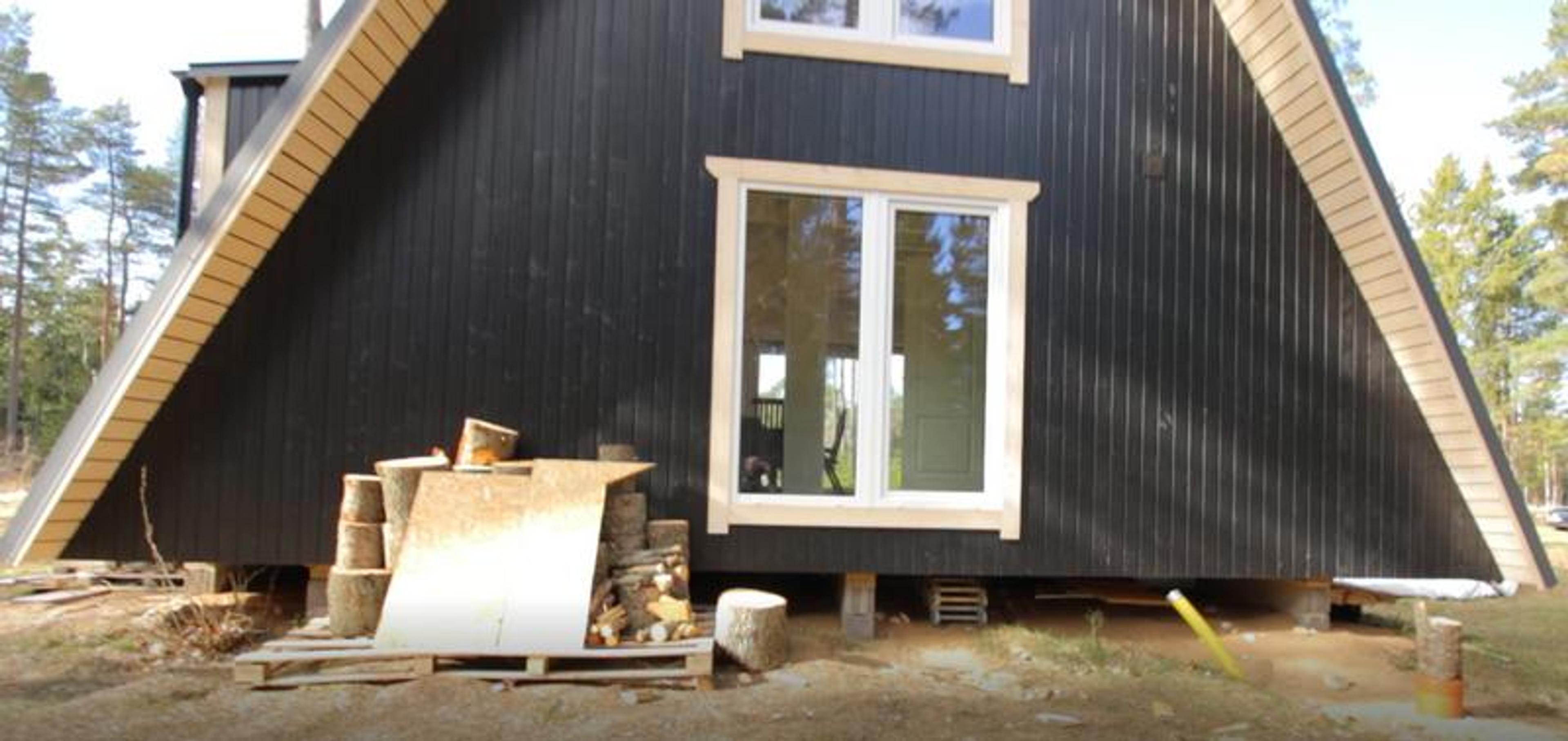 The side profile of a black-sided Trio 57 A-frame house shows the structure elevated on a concrete block foundation with firewood stacked in the foreground.