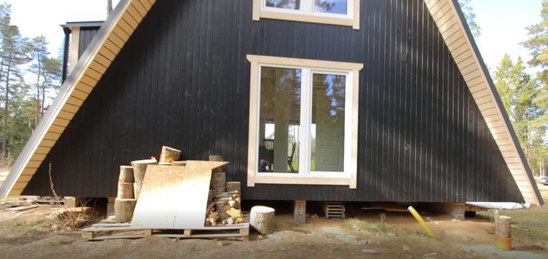The side profile of a black-sided Trio 57 A-frame house shows the structure elevated on a concrete block foundation with firewood stacked in the foreground.