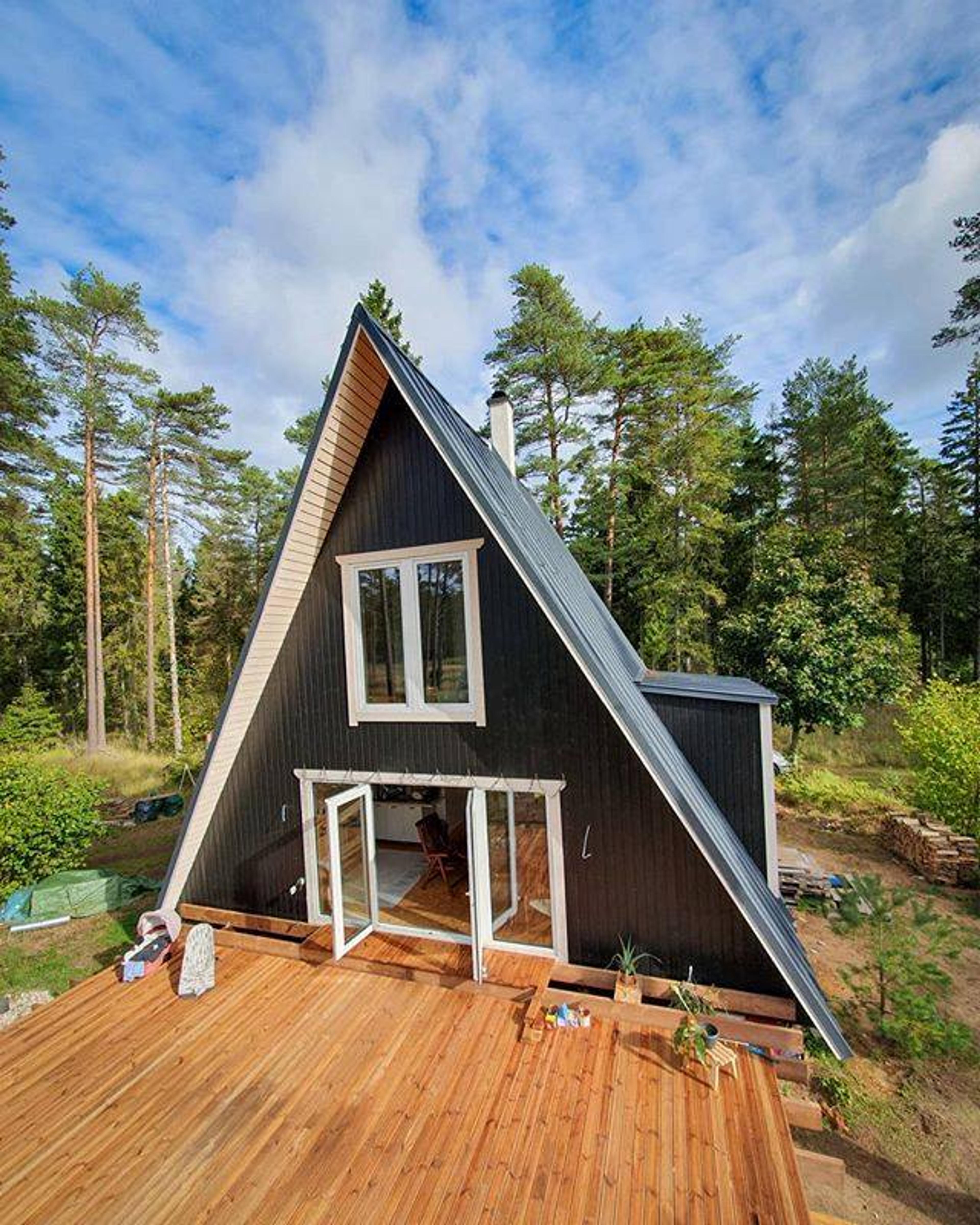 A high-angle shot shows a black A-frame cabin with white-trimmed windows and open double doors leading onto a large, newly constructed wooden deck surrounded by a lush pine forest.