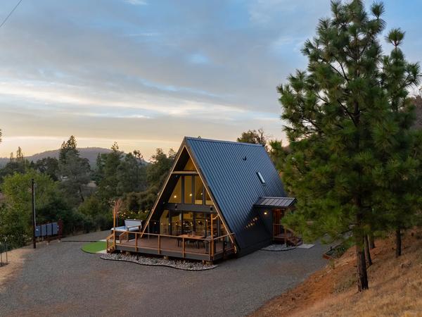 A modern A-frame house with a dark metal roof and warm interior lighting features a spacious wooden deck nestled in a hilly, pine-forested landscape during twilight.