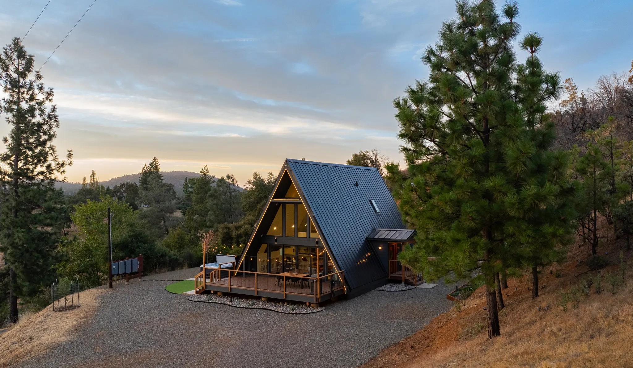 A modern A-frame house with a dark metal roof and warm interior lighting features a spacious wooden deck nestled in a hilly, pine-forested landscape during twilight.