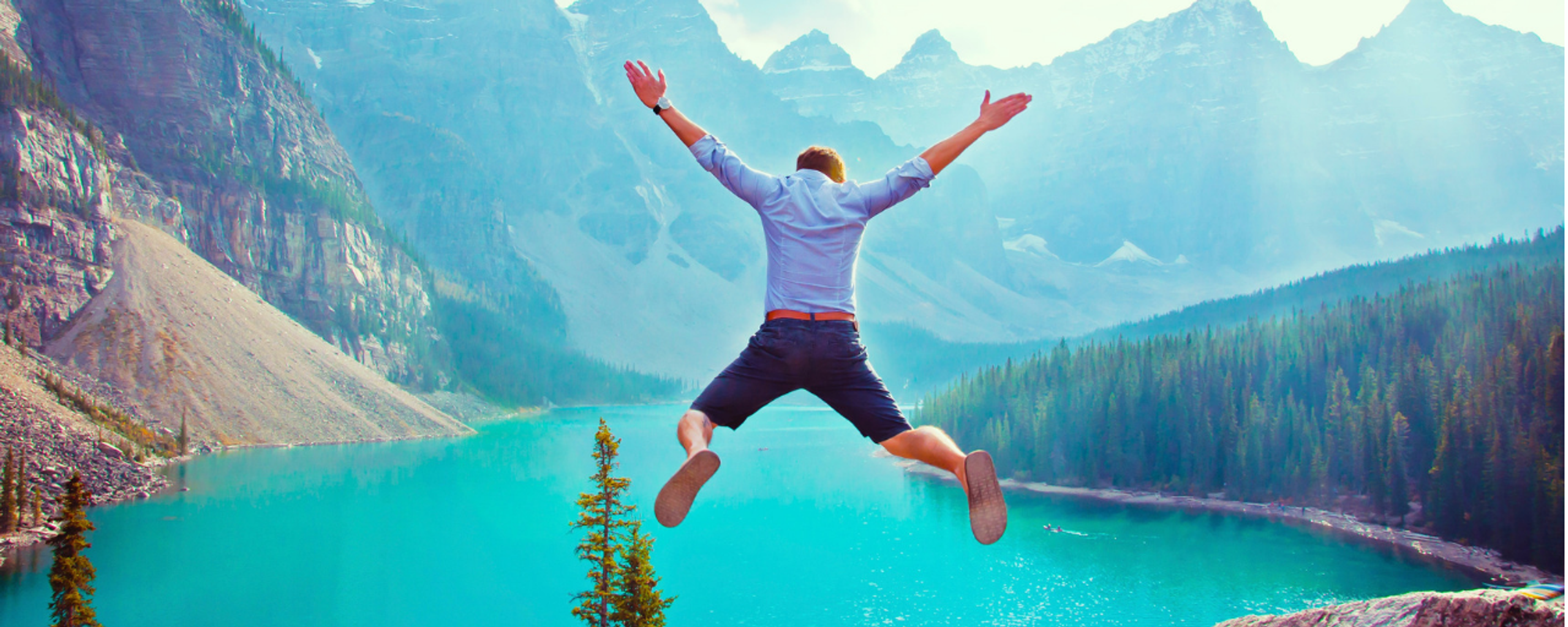 A man jumps joyfully into the air with outstretched arms, overlooking a stunning turquoise alpine lake surrounded by evergreen forests and sunlit, snow-capped mountains.