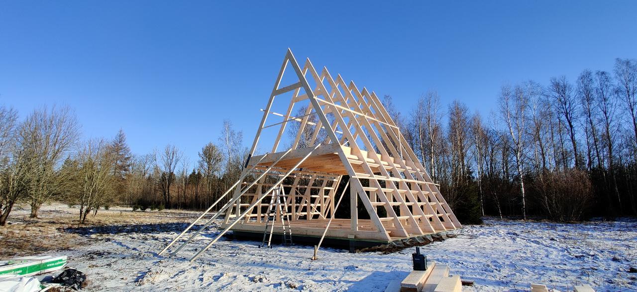 The wooden structural framework of an A-frame house continues to take shape in a snowy field, with additional horizontal purlins and floor joists installed.