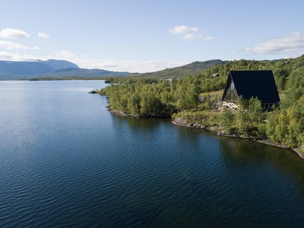 An aerial view shows a dark A-frame house situated on a wooded shoreline overlooking a vast, calm blue lake with mountains in the distance.