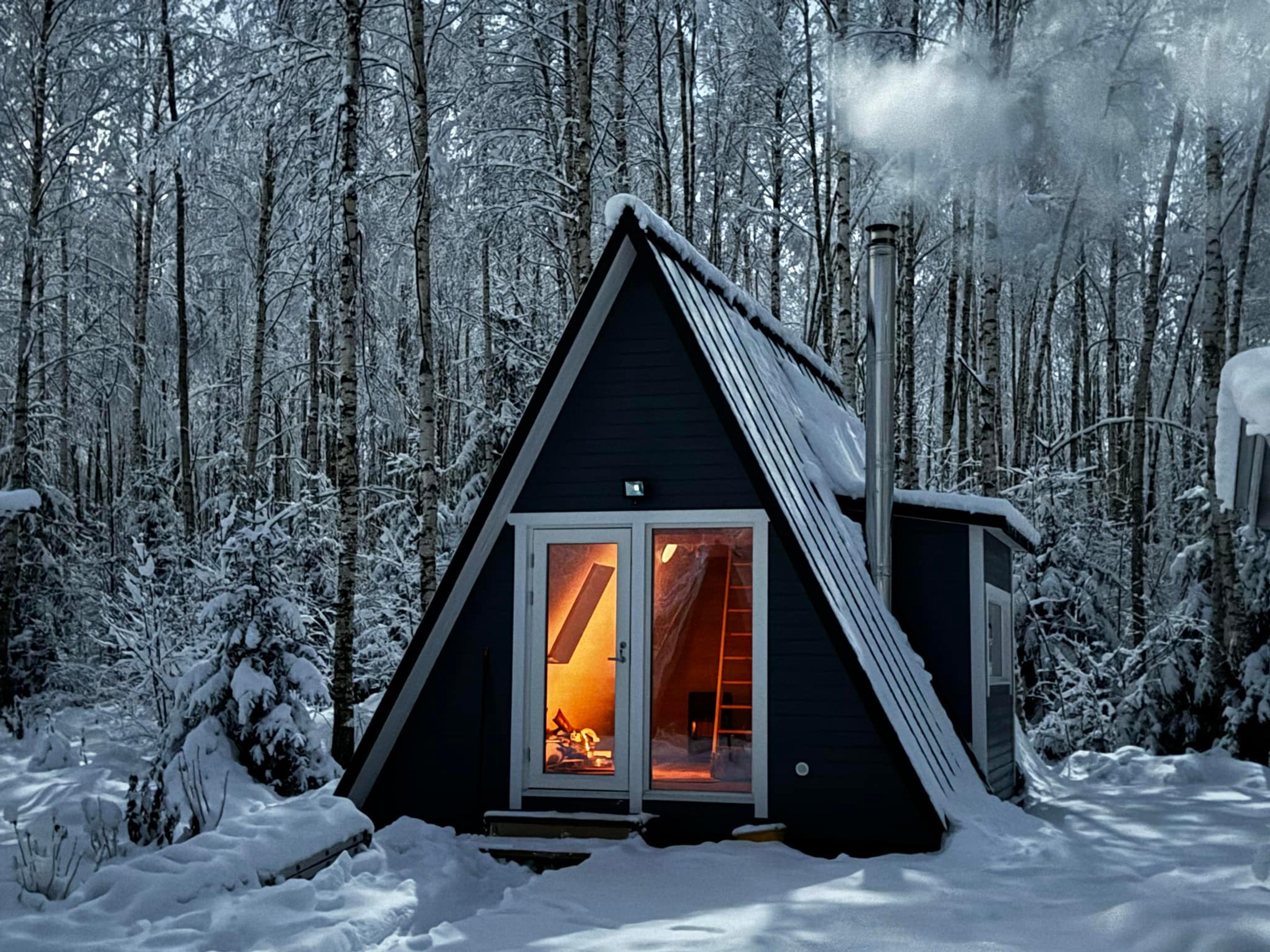 A small, warmly lit A-frame cabin stands in a snow-covered forest at twilight, with smoke rising from its metal chimney.