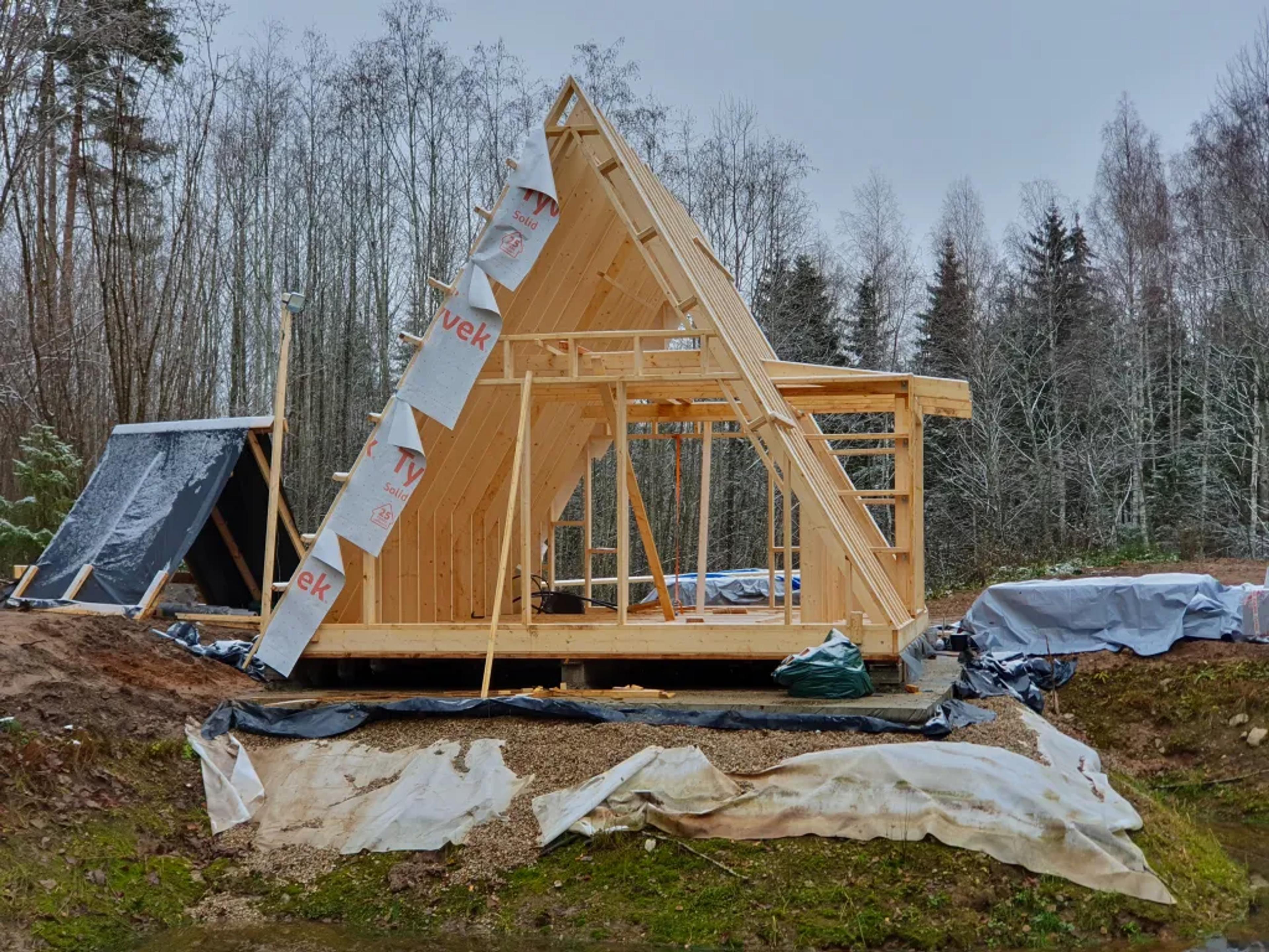 An A-frame house under construction shows the timber framing partially covered with weather-resistant house wrap in a wooded setting.