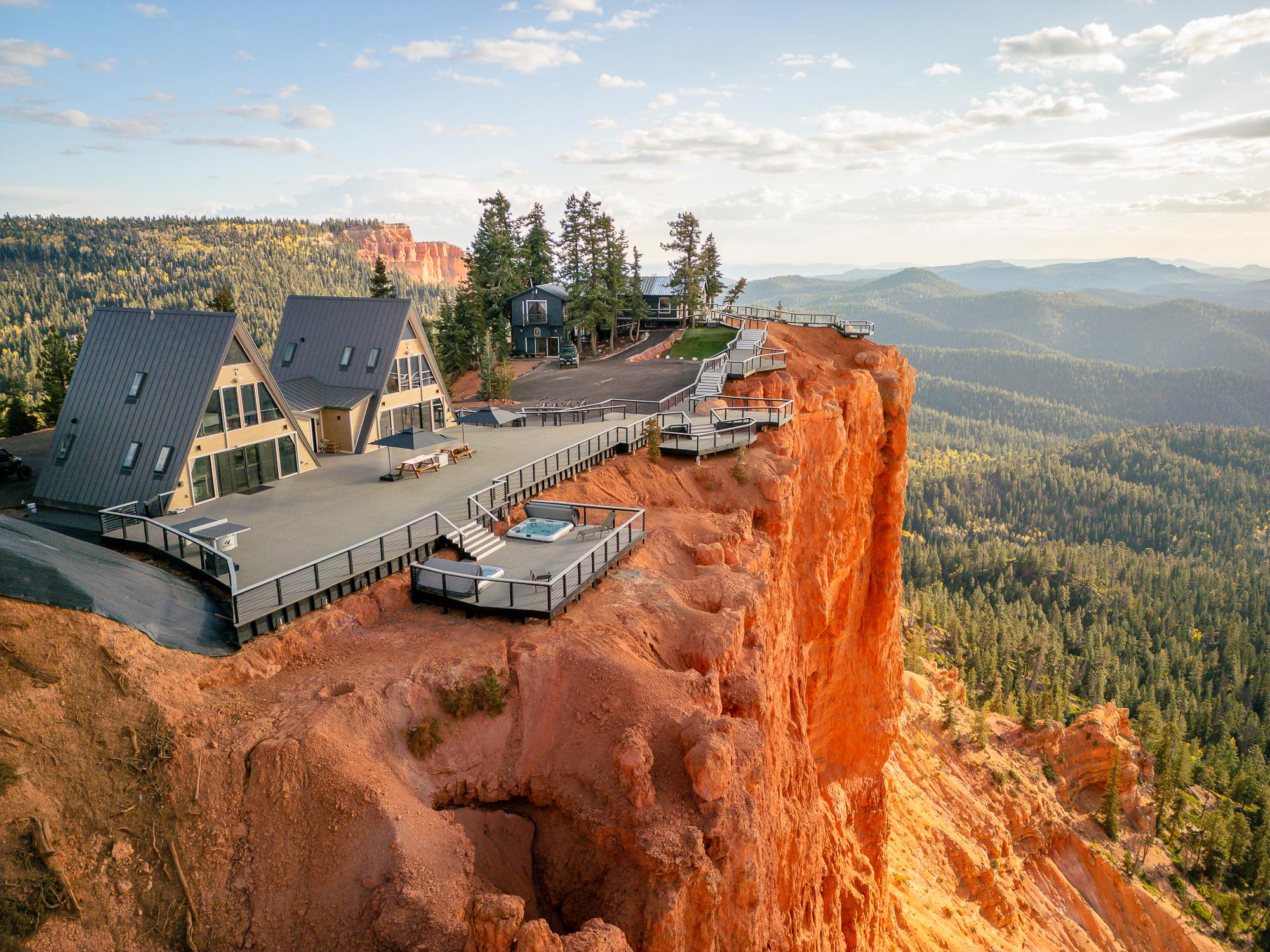An aerial perspective of 'Zen Nest,' a stunning A-frame cabin retreat perched dramatically on the edge of a high, red-rock cliff. The cabin features multiple connected A-frame structures with dark metal roofs, an expansive wooden deck, a hot tub, and sweeping views of a vast pine forest and mountainous valley below.
