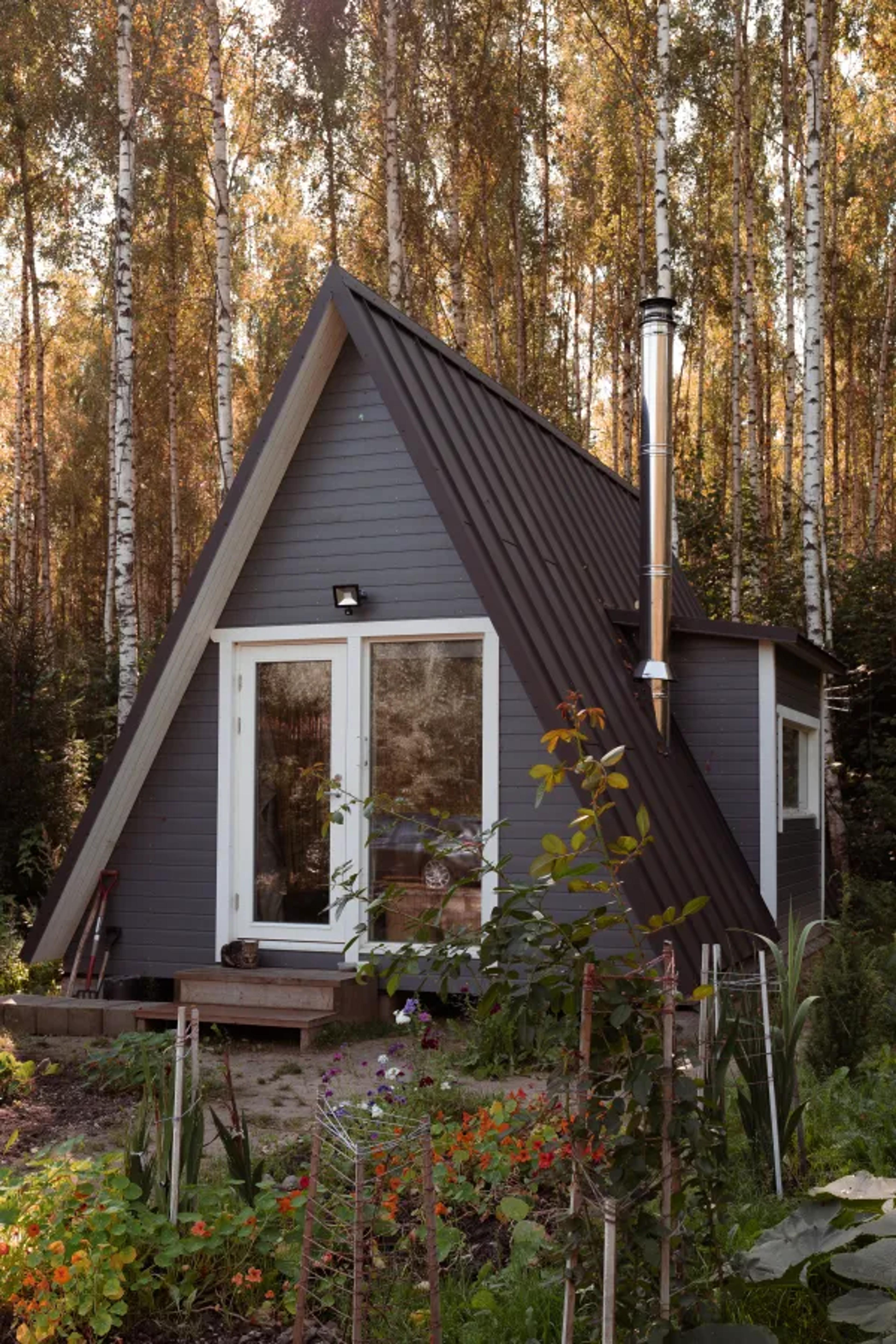 A dark grey A-frame cabin with white door trim and a tall metal chimney sits in a lush garden clearing surrounded by tall, slender trees.