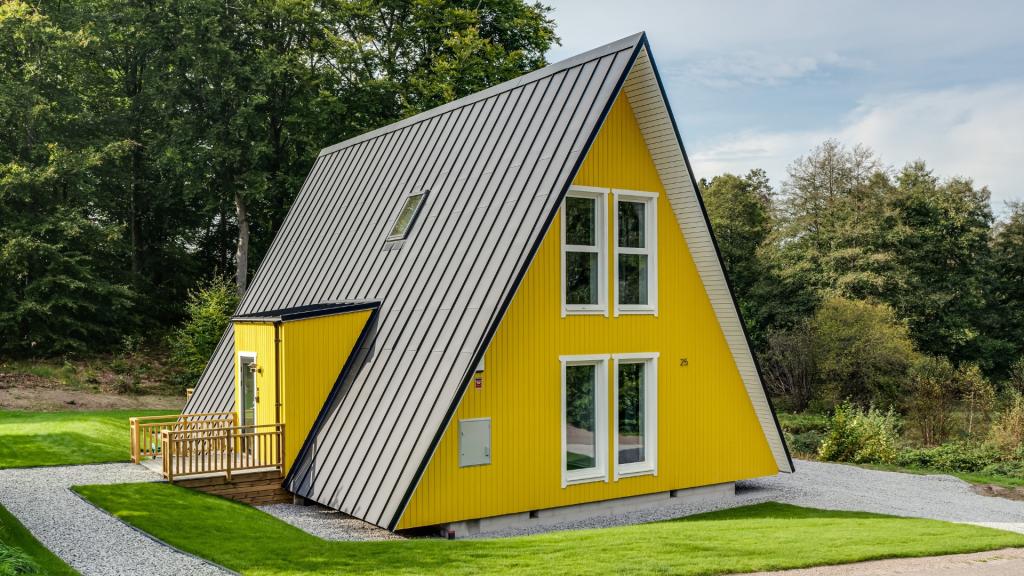 A finished Avrame Trio 120 house features vibrant yellow siding, a dark metal roof, and a small wooden porch, all set against a lush green lawn and forest backdrop.