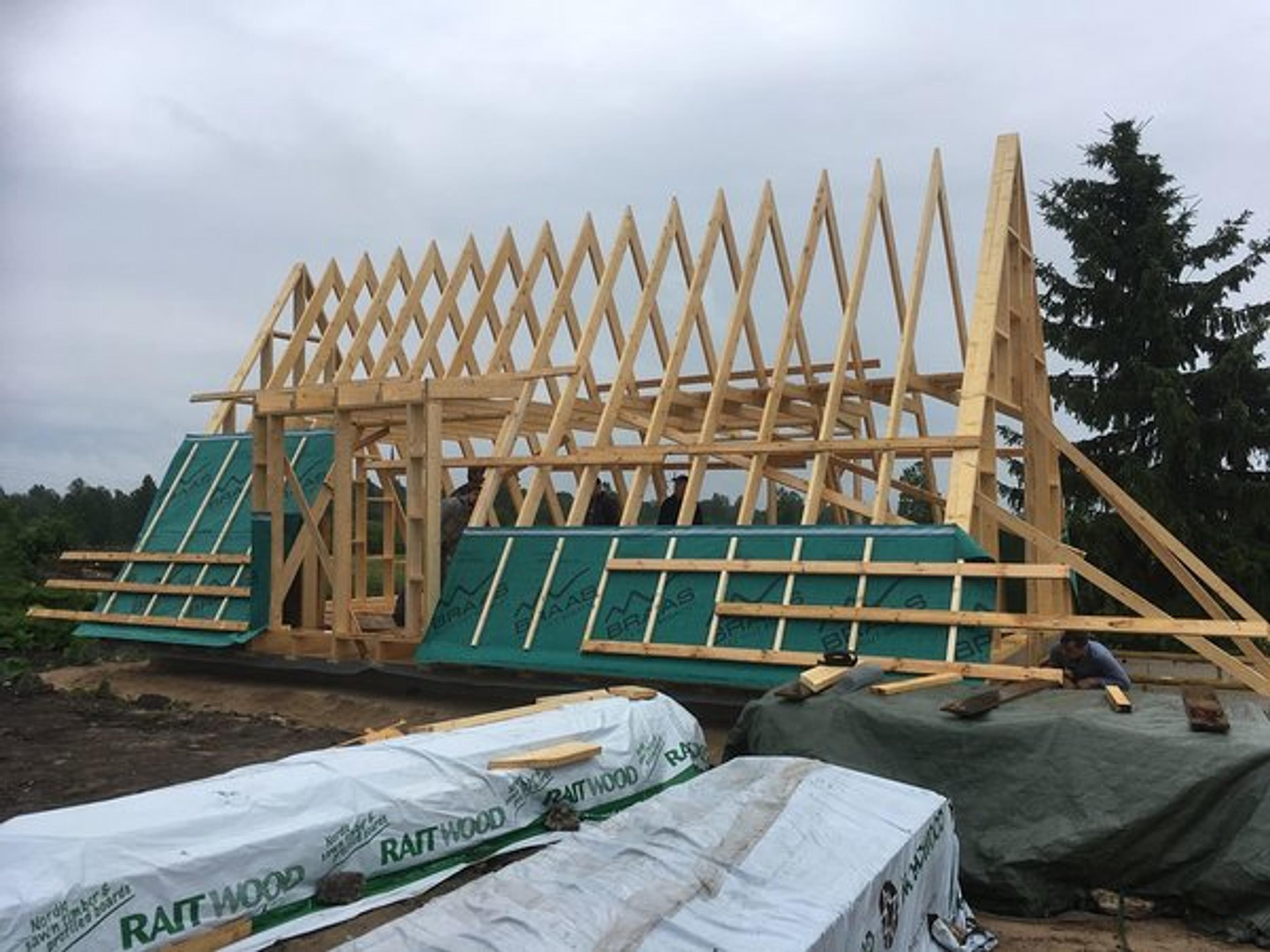 A wooden A-frame skeletal structure is under construction, with green weather-resistant membrane and horizontal battens partially installed over the lower sections of the trusses.
