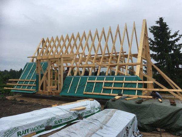 A wooden A-frame skeletal structure is under construction, with green weather-resistant membrane and horizontal battens partially installed over the lower sections of the trusses.
