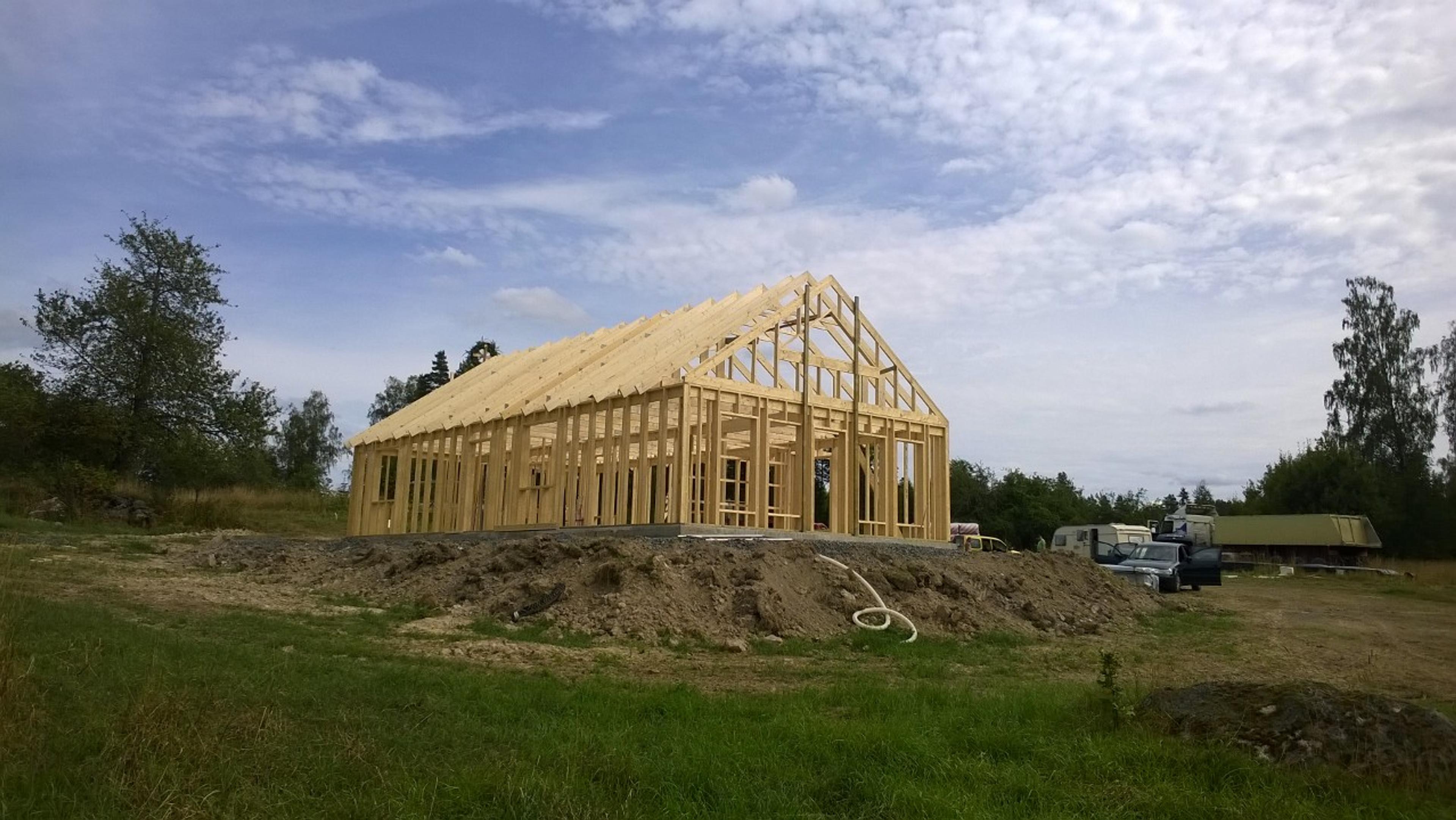 The wooden wall studs and roof trusses of a large, rectangular gabled house stand partially assembled on a dirt construction site under a blue sky.