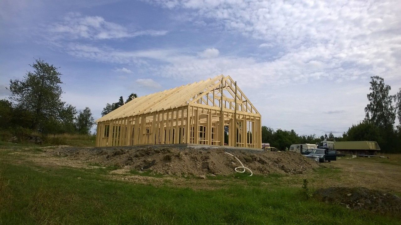 The wooden wall studs and roof trusses of a large, rectangular gabled house stand partially assembled on a dirt construction site under a blue sky.