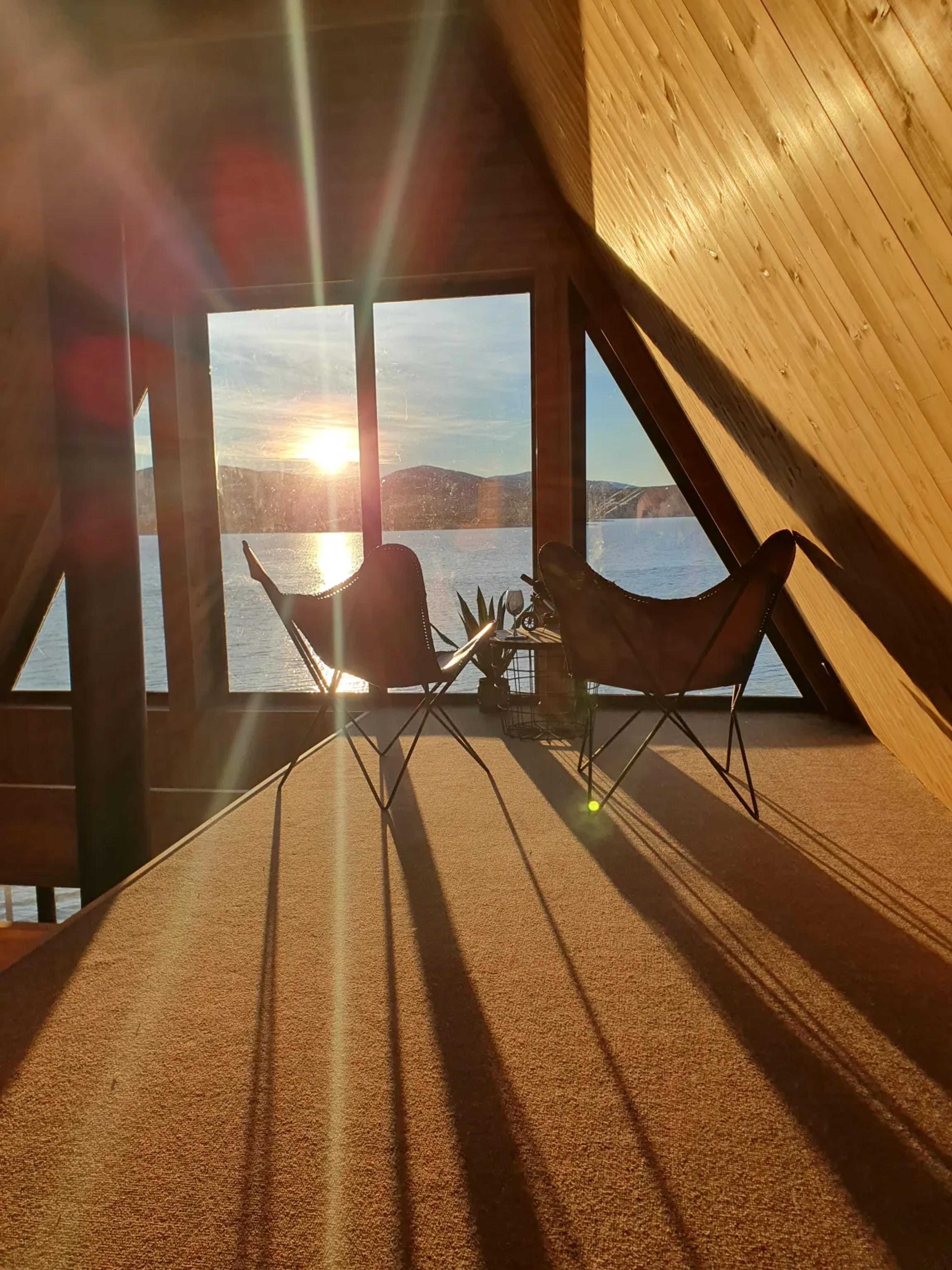 Two butterfly chairs are positioned on the carpeted loft of an A-frame house, facing a large window that overlooks a sunset reflecting on a lake.