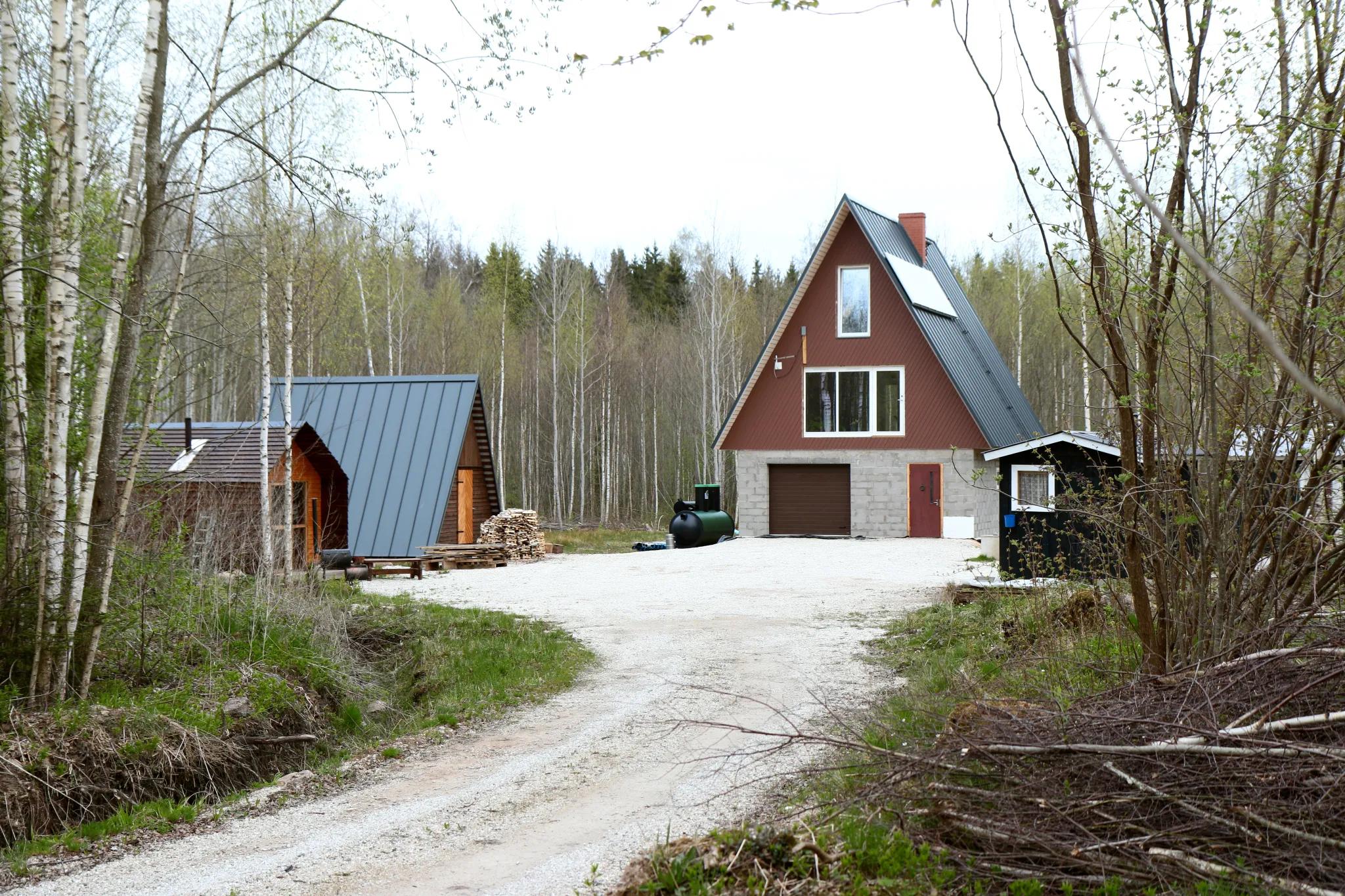 A gravel driveway leads toward a rural property featuring a large A-frame house with a concrete block garage base and a smaller A-frame cabin, both set against a dense forest backdrop.