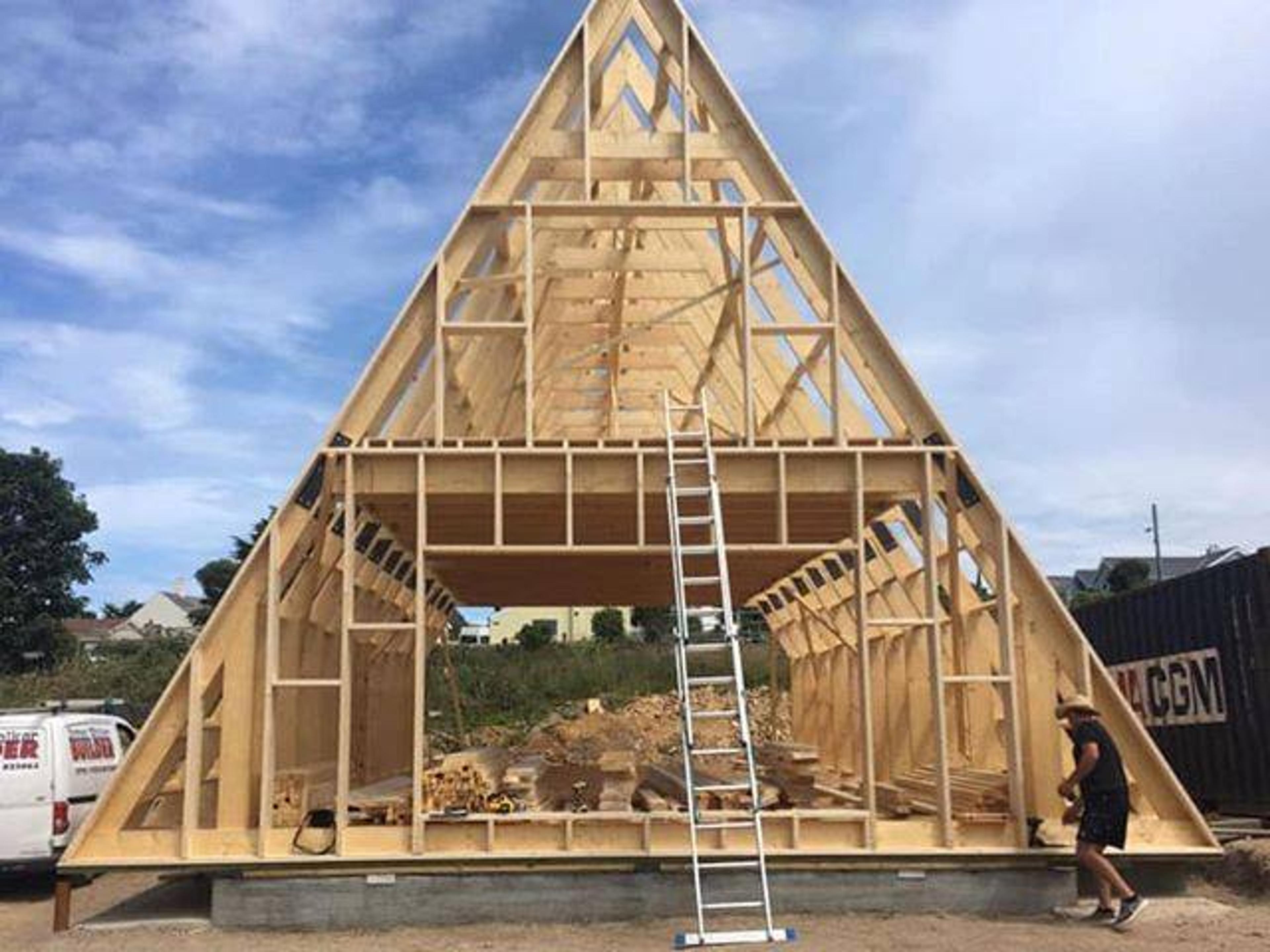 The wooden skeletal frame of a multi-story A-frame house is shown under construction with a tall ladder leaning against the second-story floor joists.