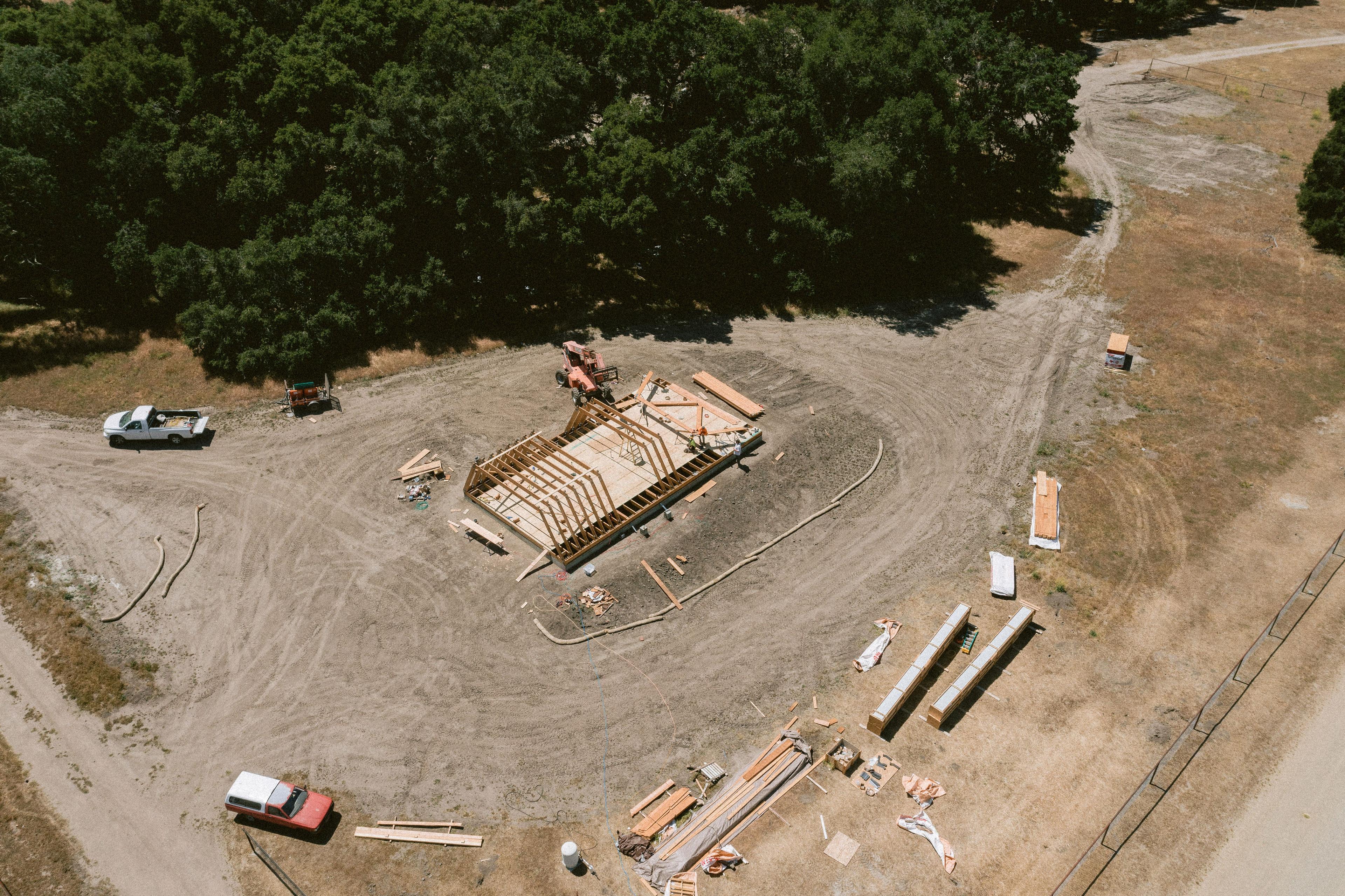 An aerial drone view of an active house construction project showing the precise assembly of an Avrame A-frame self build kit. The image highlights a clean, organized job site with the floor joists and initial triangular rafters being raised in a rural setting, demonstrating minimal material waste.