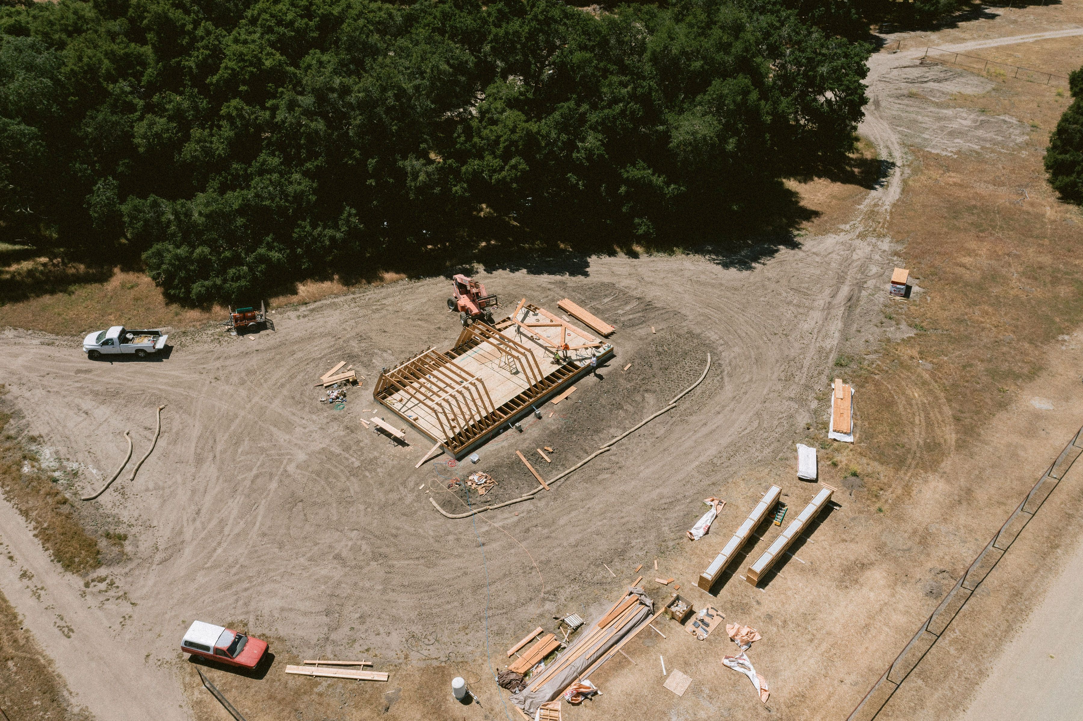 An aerial drone view of an active house construction project showing the precise assembly of an Avrame A-frame self build kit. The image highlights a clean, organized job site with the floor joists and initial triangular rafters being raised in a rural setting, demonstrating minimal material waste.