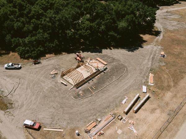 An aerial drone view of an active house construction project showing the precise assembly of an Avrame A-frame self build kit. The image highlights a clean, organized job site with the floor joists and initial triangular rafters being raised in a rural setting, demonstrating minimal material waste.