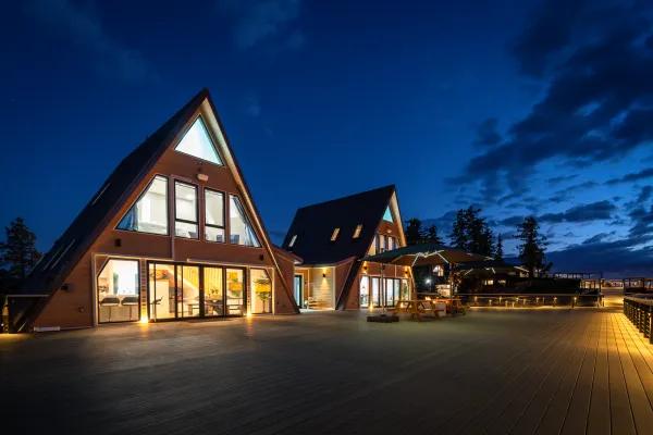 Two modern, glowingly lit A-frame cabins sit on an expansive wooden terrace at night, complete with outdoor seating and umbrellas under a deep blue evening sky.