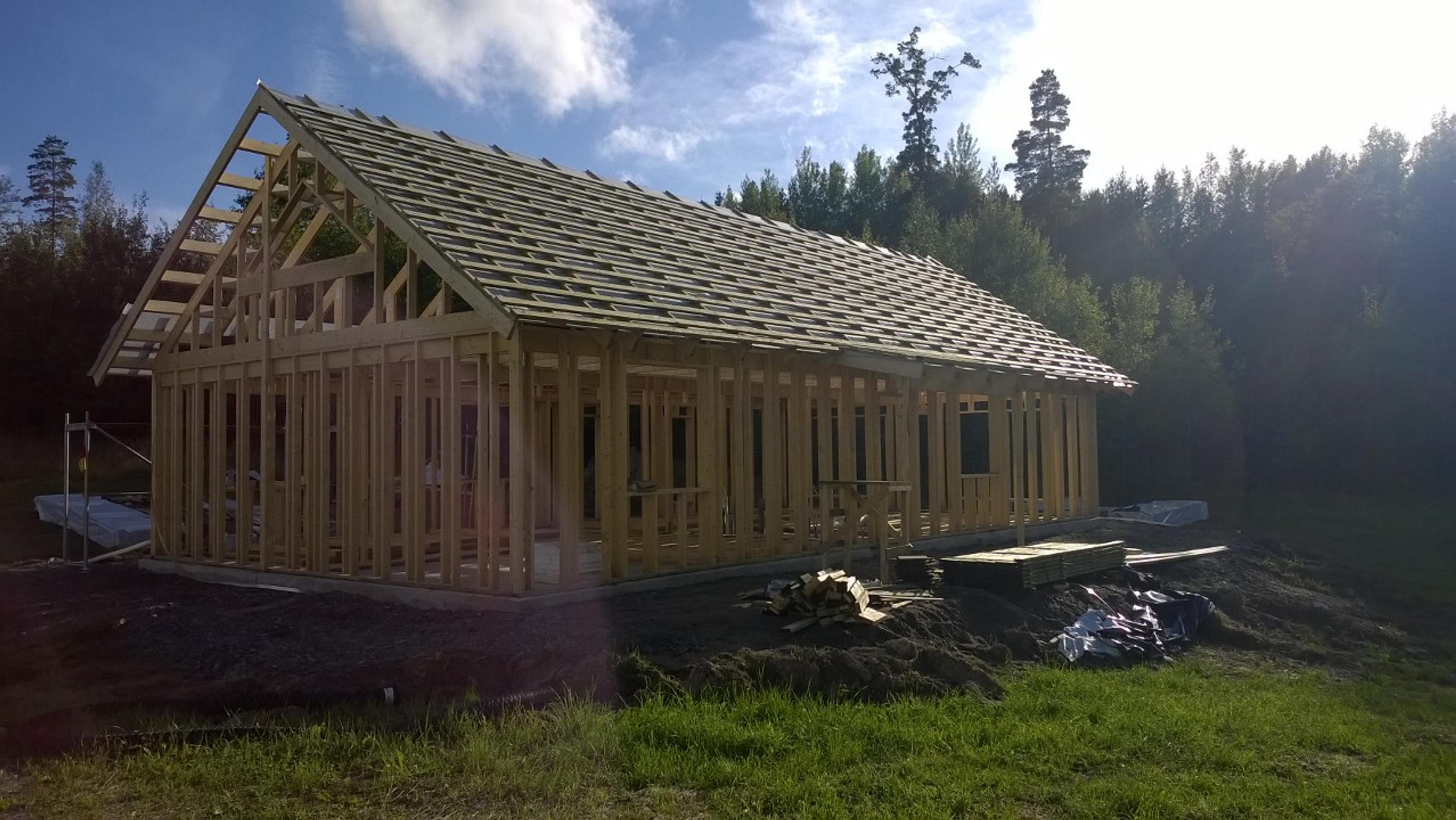 The wooden frame of a gabled house is shown with roof battens installed over the trusses against a backdrop of dense forest.