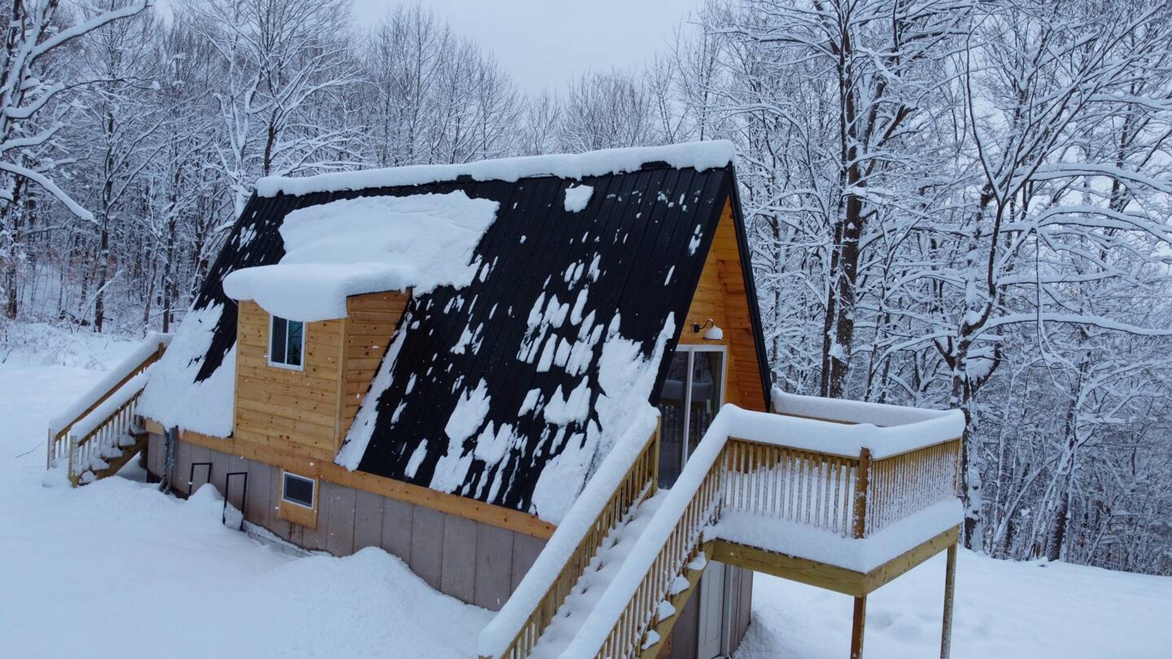 An elevated view shows a wooden A-frame cabin with a snow-dusted black metal roof and a large wraparound deck, set against a dense, snow-covered forest.