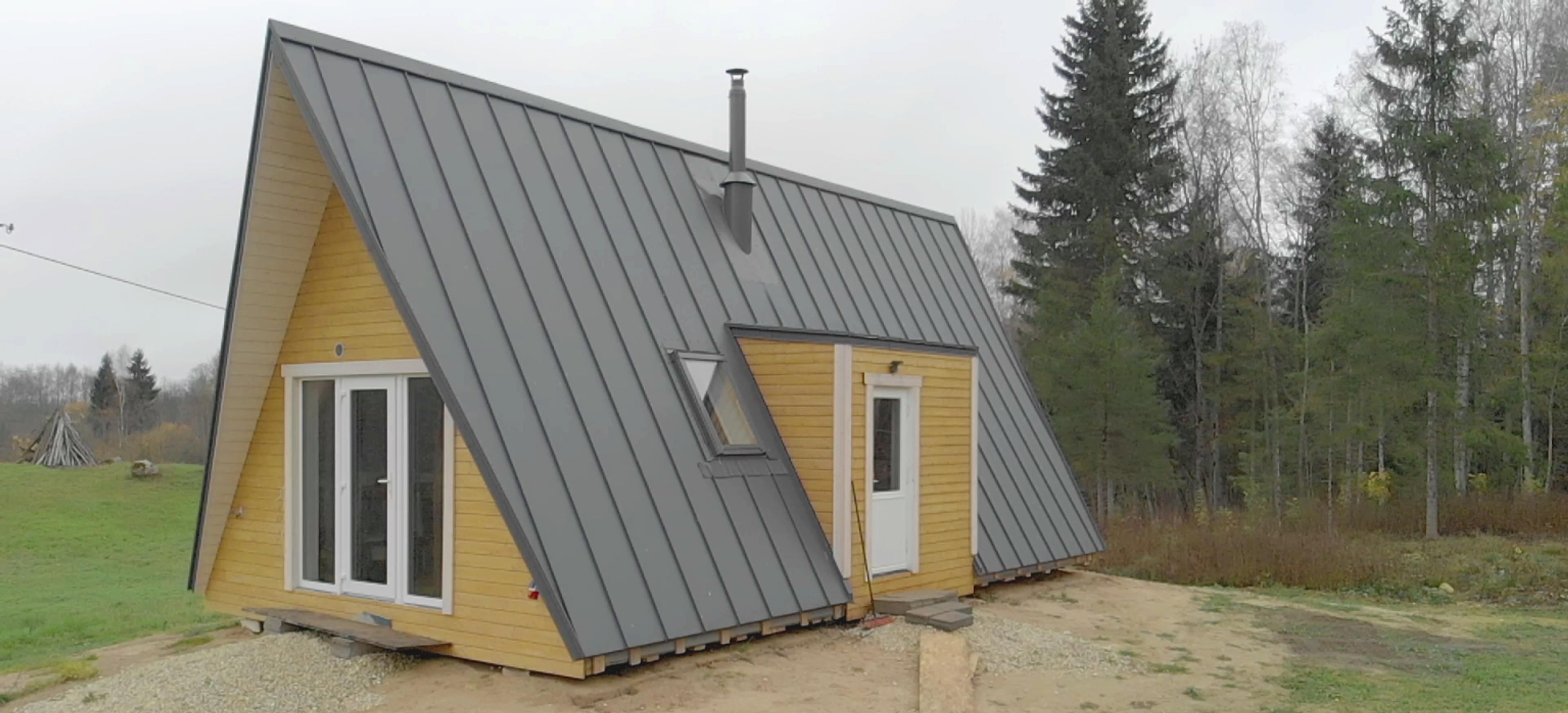 A yellow A-frame house features a large, grey standing-seam metal roof with an integrated side dormer and a central chimney flue.